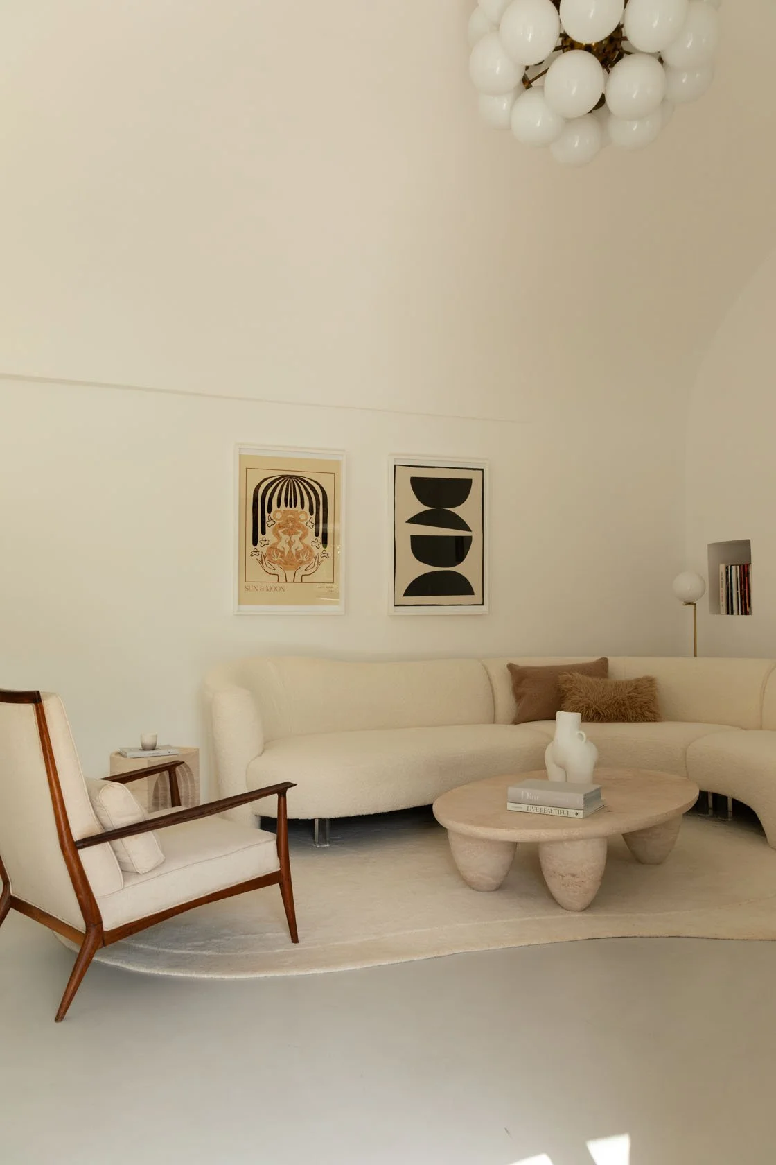 Living room with a cream-colored curved sofa, a wooden armchair with white cushions, a round beige coffee table with books and a white vase, framed abstract art on the wall, a white spherical lamp on a stand, and a decorative ceiling light made of wh
