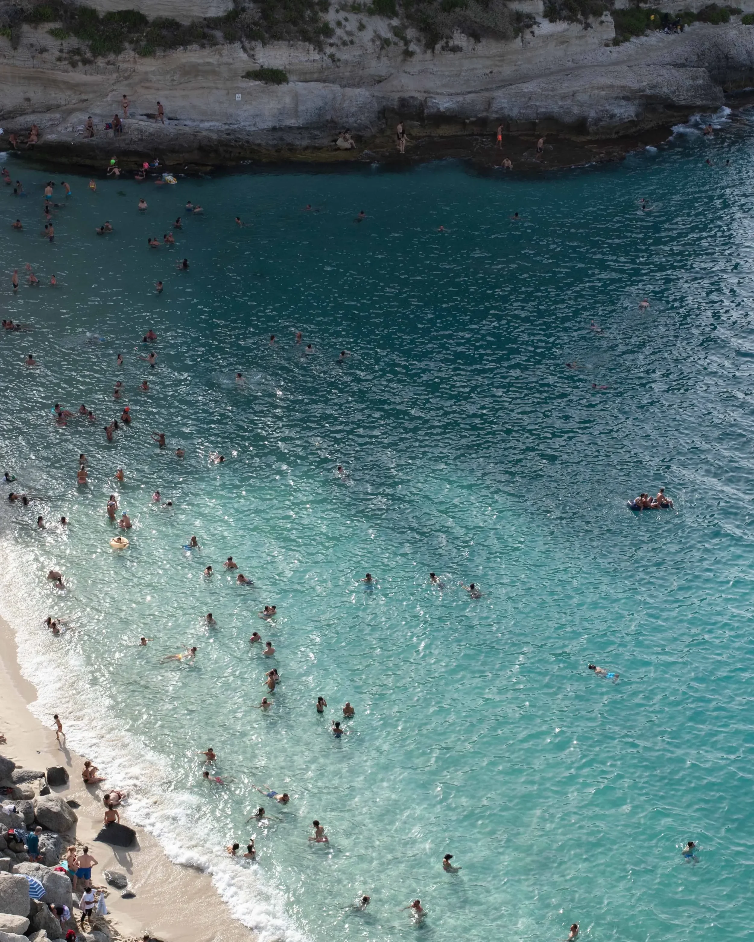 People swimming and relaxing along a rocky beach with clear blue water and cliffs at the background.