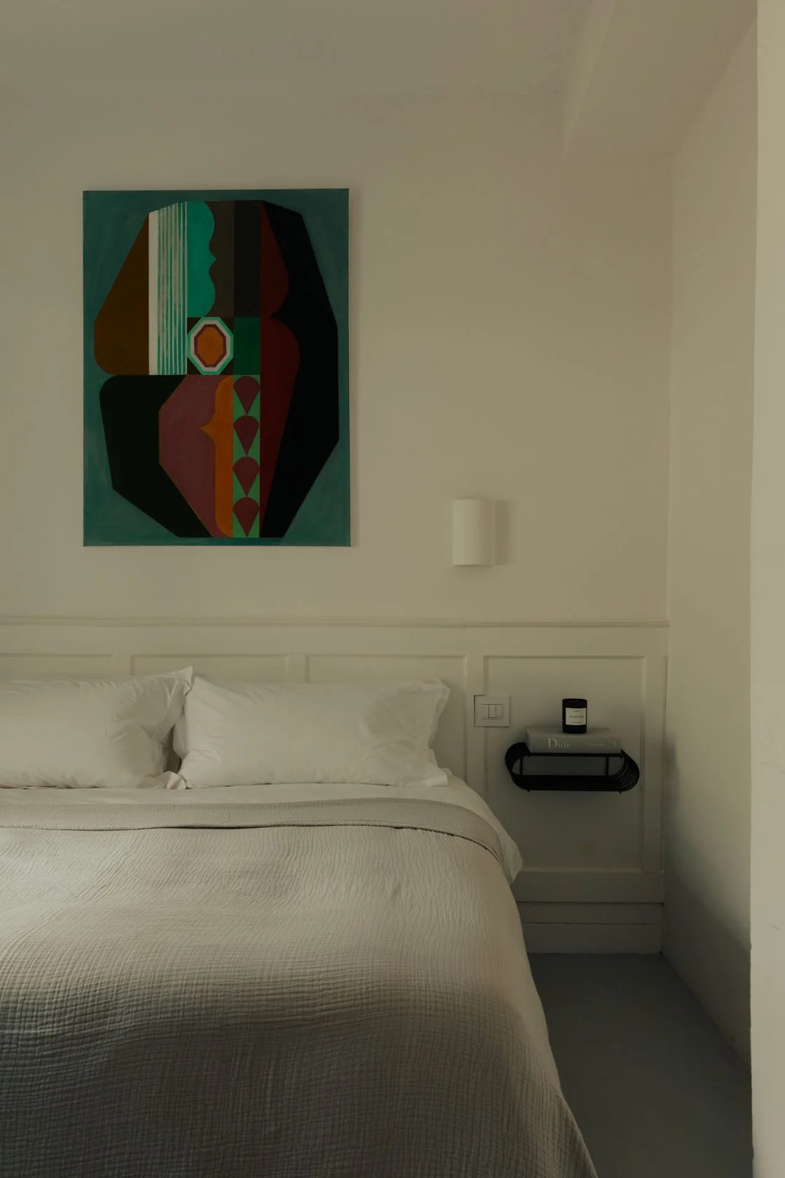 A cozy hotel room with a neatly made bed, white pillows, and a beige bedspread. A colorful abstract painting hangs above the bed. To the side, there is a small black wall shelf with a book titled 'Dior' and a black candle.