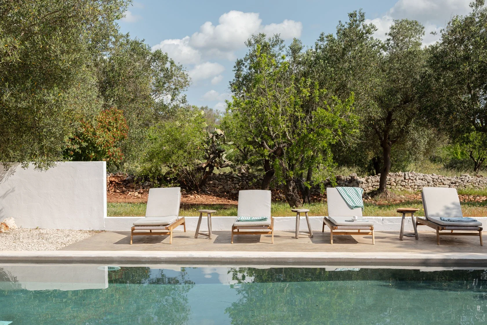 Row of lounge chairs with towels by a swimming pool, surrounded by trees and a stone wall in the background, under a partly cloudy sky.