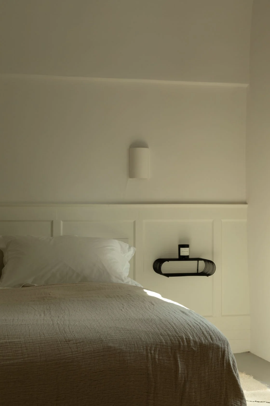 A bedroom with a white bed, white pillow, beige bedspread, and a black wall shelf holding a black container, against a plain cream-colored wall with a white wall-mounted light fixture.