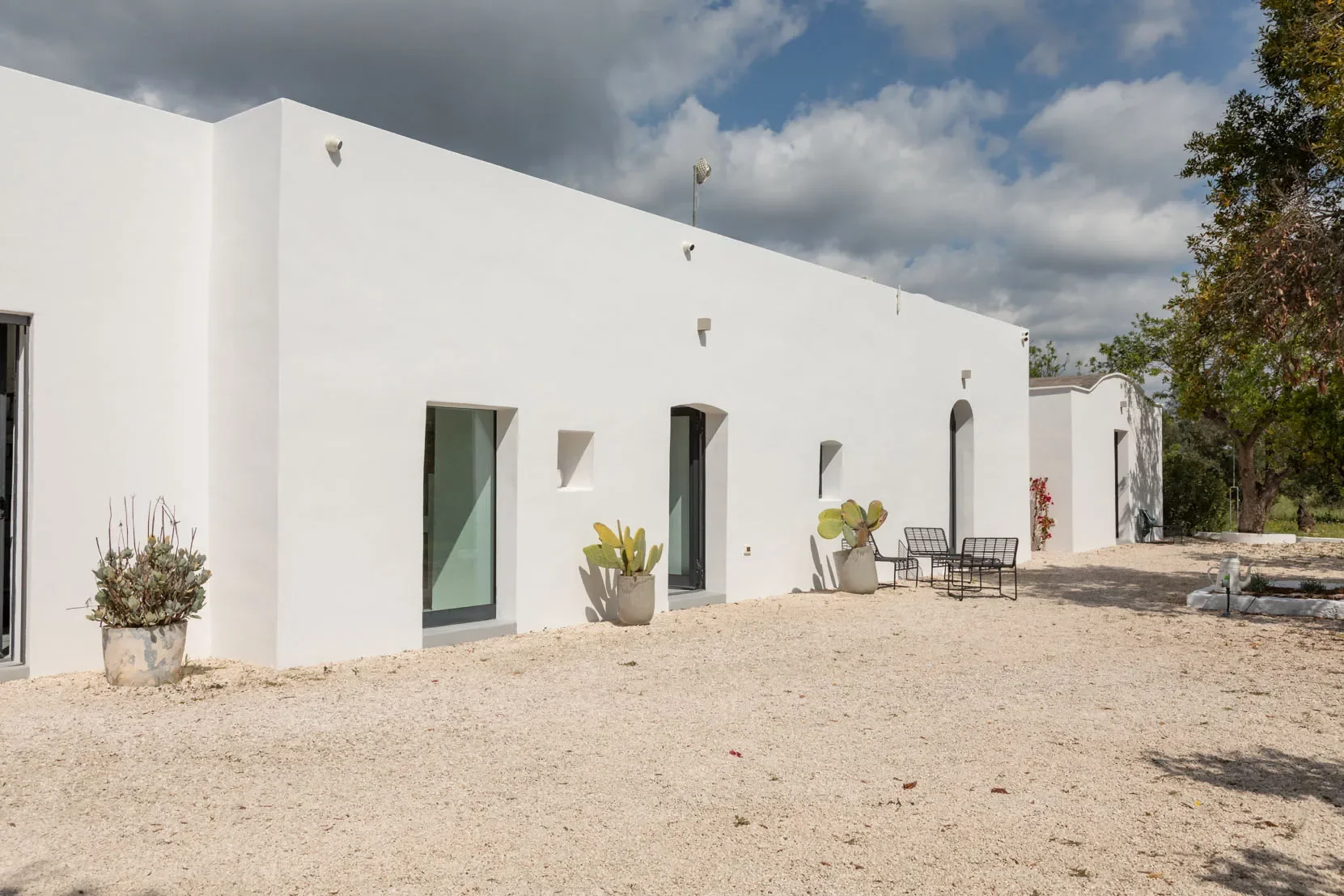 A white modern building with multiple windows, potted plants, and outdoor seating on a gravel ground under a partly cloudy sky.
