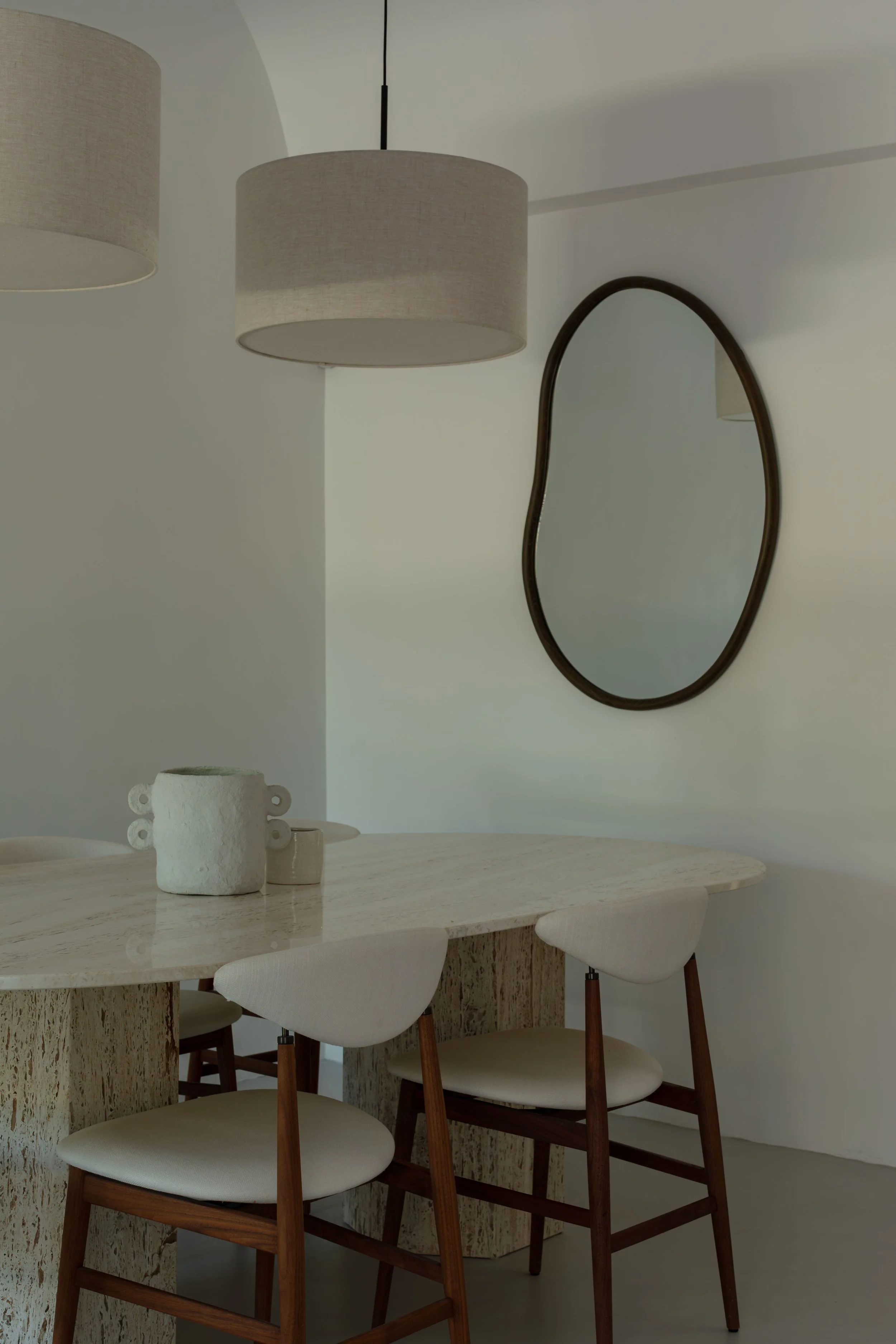 Interior dining area with a marble table, three white chairs with wooden legs, a wall mirror with an irregular shape, layered pendant lamps, and decorative jars on the table.