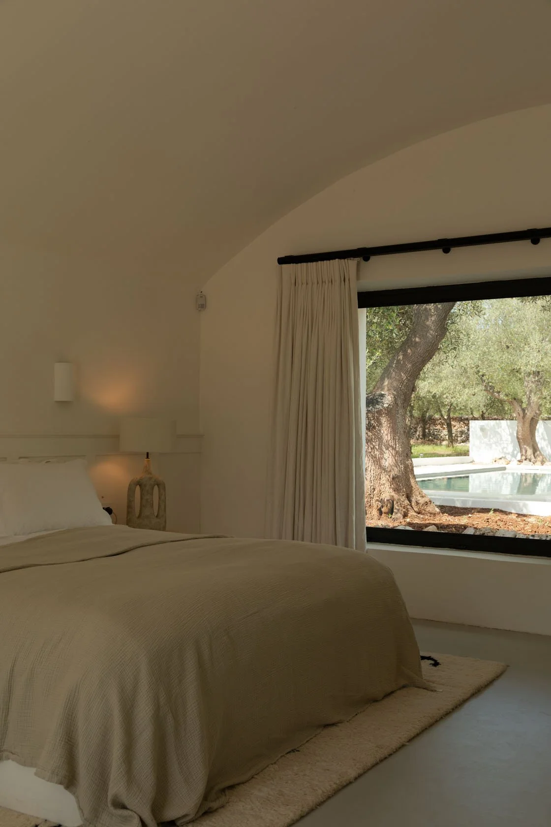 A cozy bedroom with a beige bedspread, a modern bedside lamp on a sculptural nightstand, and a large window with white curtains showing an outdoor view of trees and a pool.