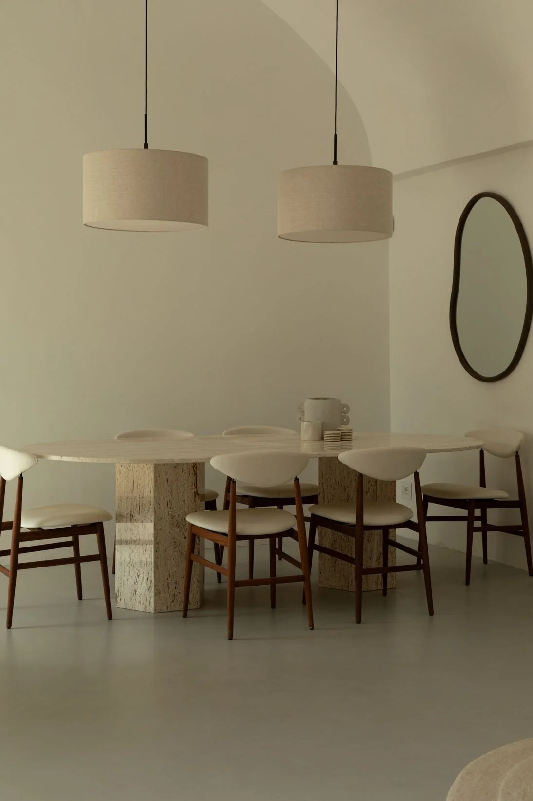 Minimalist dining room with a large oval table, six chairs with white cushions, two pendant lights, a black oval mirror on the wall, and a simple neutral color palette.