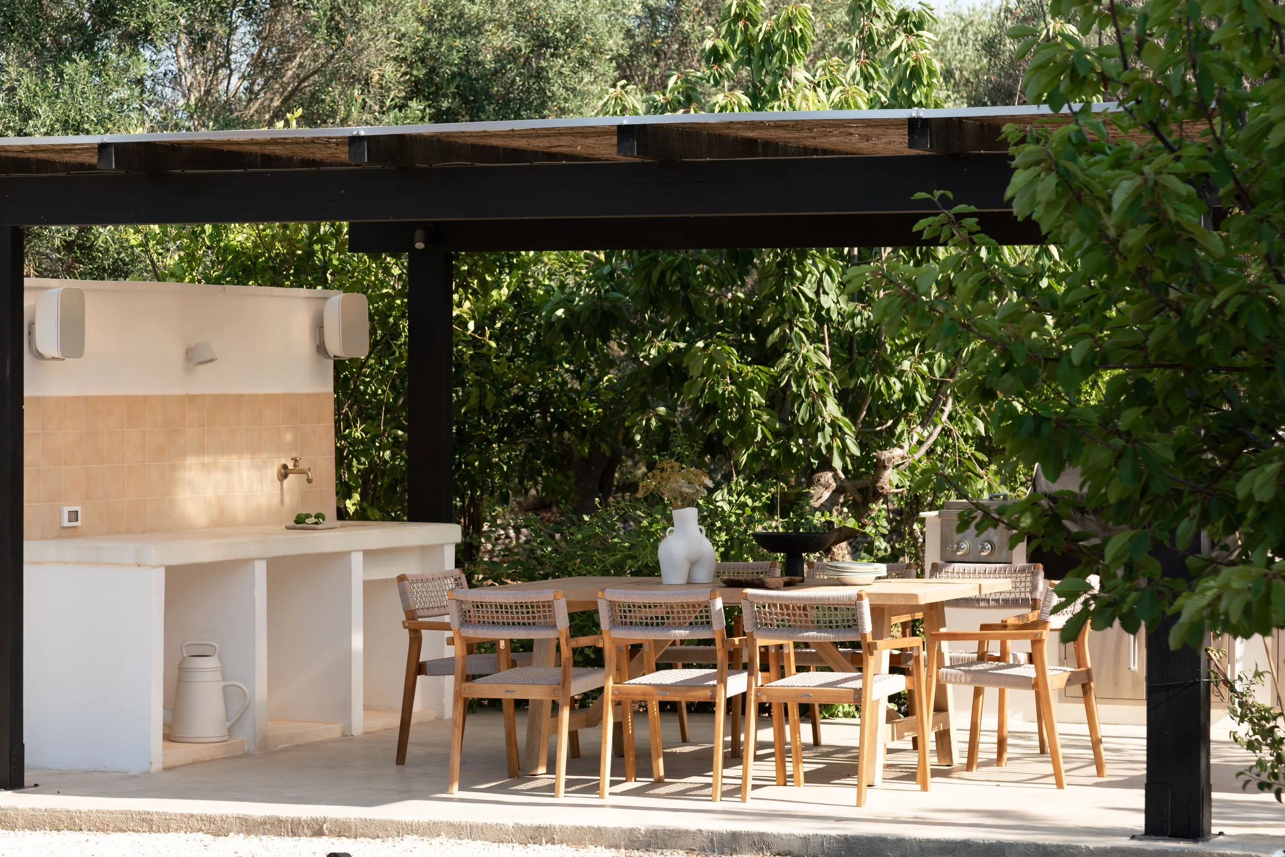 Outdoor patio with a wooden dining table and chairs, a white vase with flowers, a built-in outdoor sink with beige tile backsplash, and lush greenery in the background.