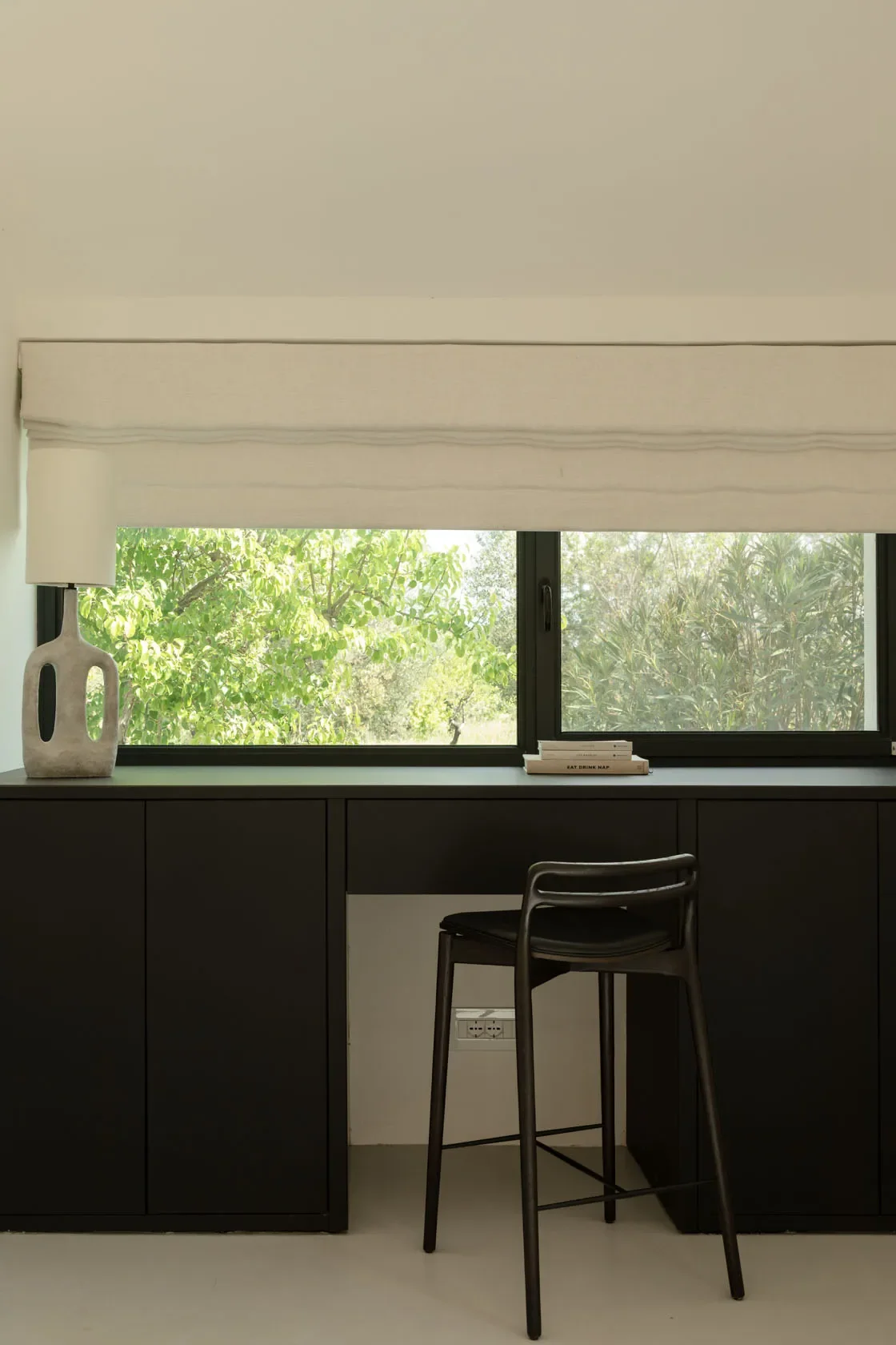 A black counter with a window behind it, showcasing trees and greenery outside. On the counter, there is a tall white vase with a sculptural design on the left and a small stack of books on the right. A black bar stool is placed in front of the count