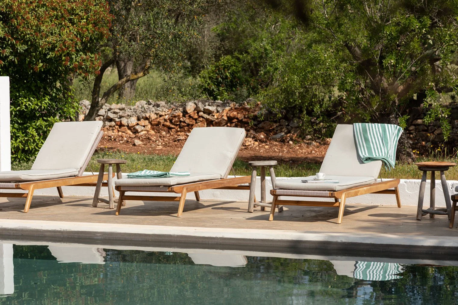 Three beige outdoor lounge chairs with wooden frames beside a swimming pool, with striped towels, mugs, and a small wooden side table on a concrete surface, surrounded by green trees and bushes.