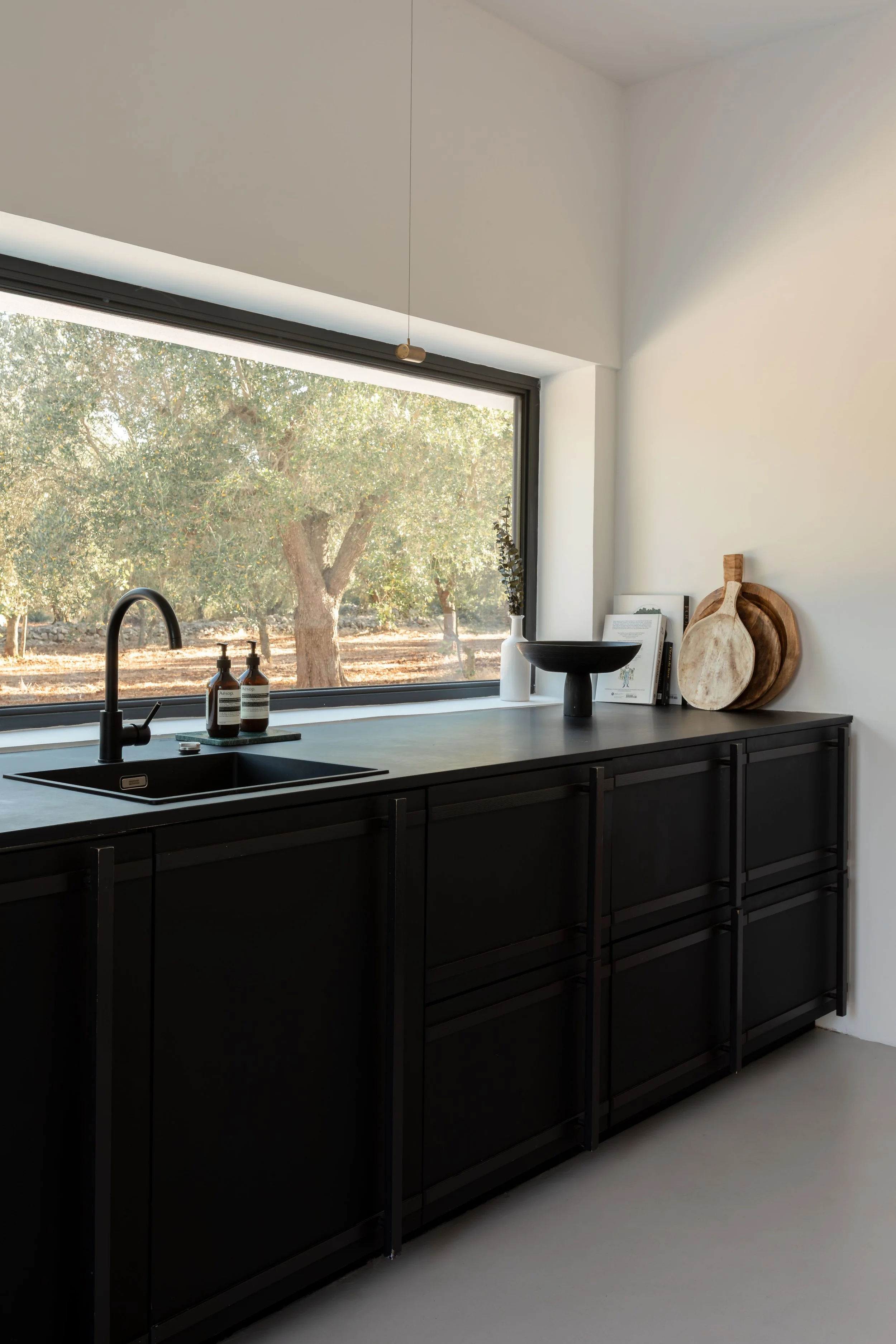 Modern minimalist kitchen with black cabinets and a large window overlooking a tree, decorated with a black bowl, a white vase with greenery, and wooden trays.