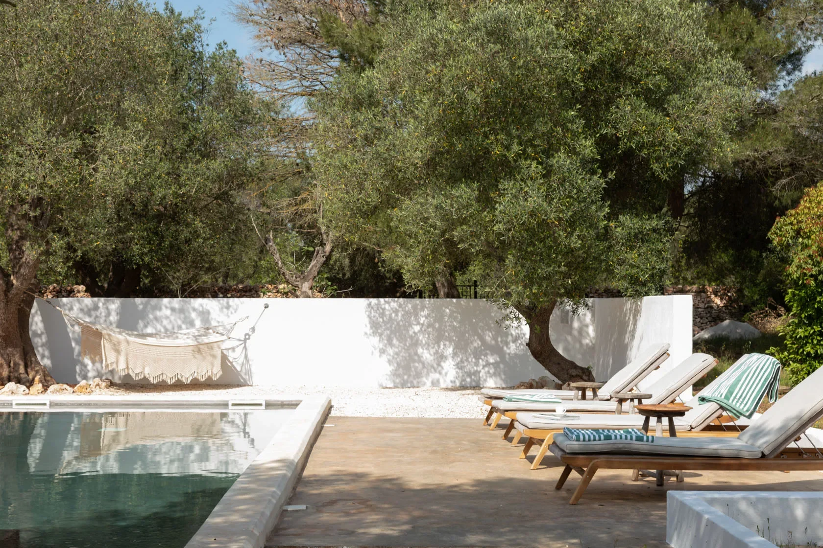 An outdoor pool area with lounge chairs, some with striped towels, a white wall with an overhanging tree, and a hammock between two trees in a sunny garden.