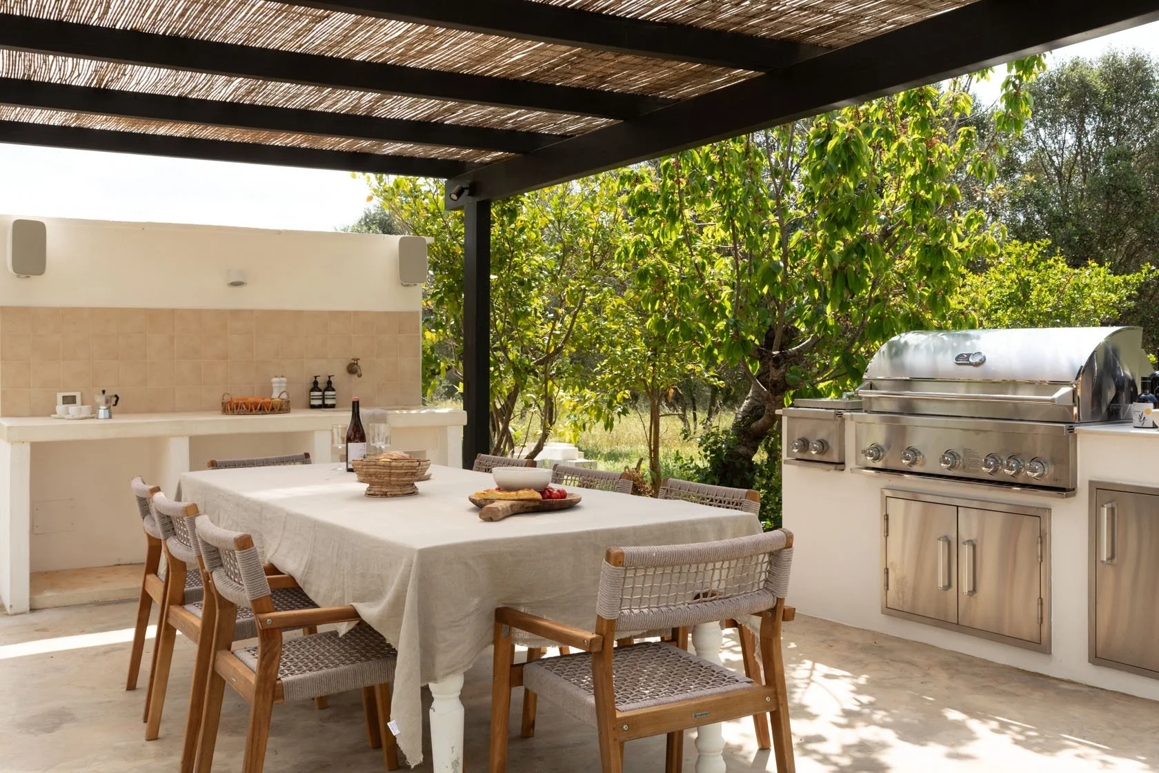 Outdoor dining area with a table covered with a beige tablecloth, surrounded by six wooden chairs, a stainless steel barbecue grill, and a backdrop of lush greenery and trees.