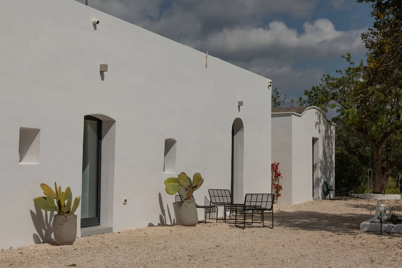 A white modern building with minimal architecture, featuring small windows and a curved roof, surrounded by cacti, outdoor furniture, and trees under a partly cloudy sky.