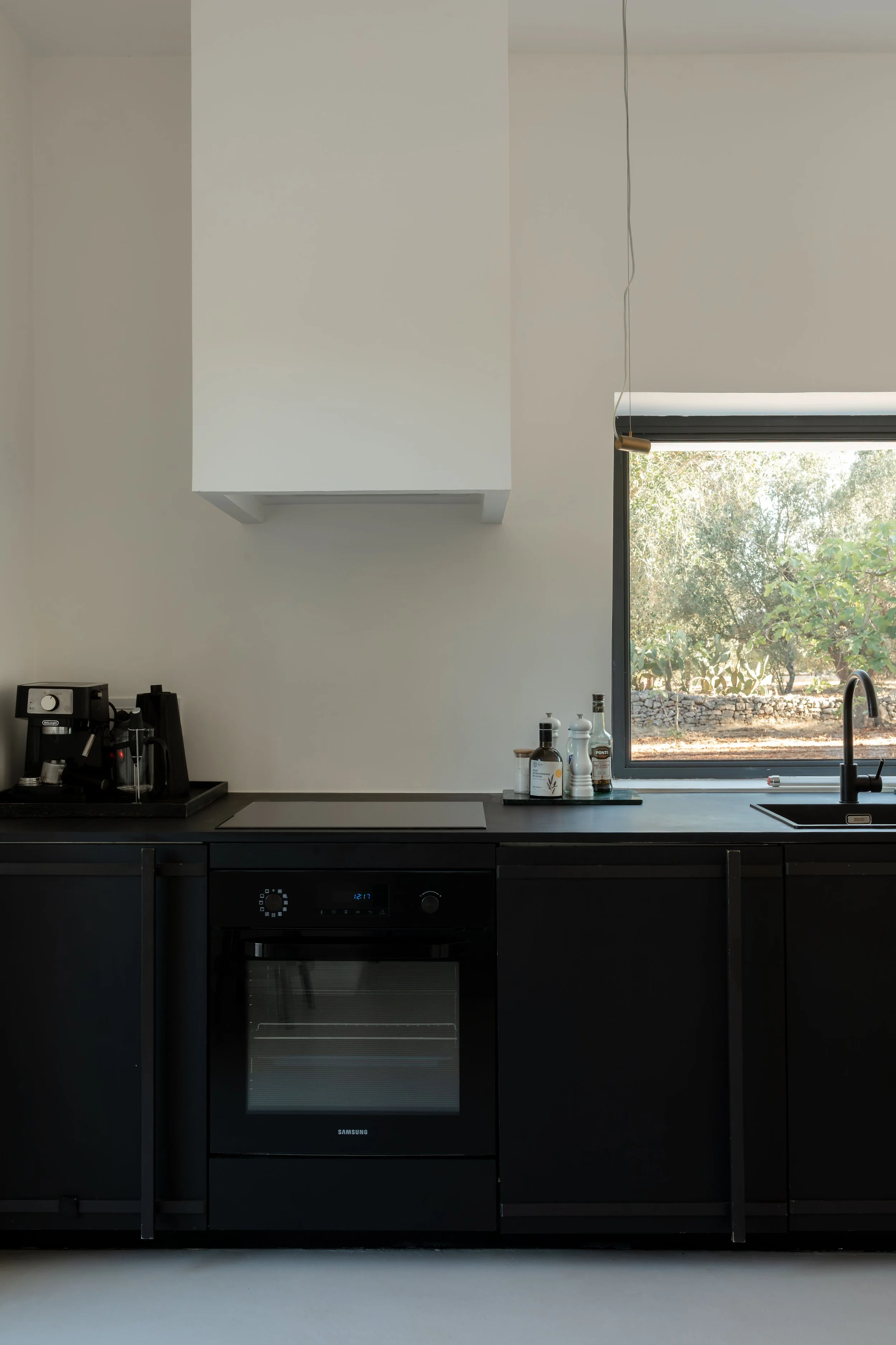 Modern kitchen with black cabinets, a window showing outdoor greenery, a built-in oven, and a stovetop, with a coffee machine and kitchen condiments on the counter.