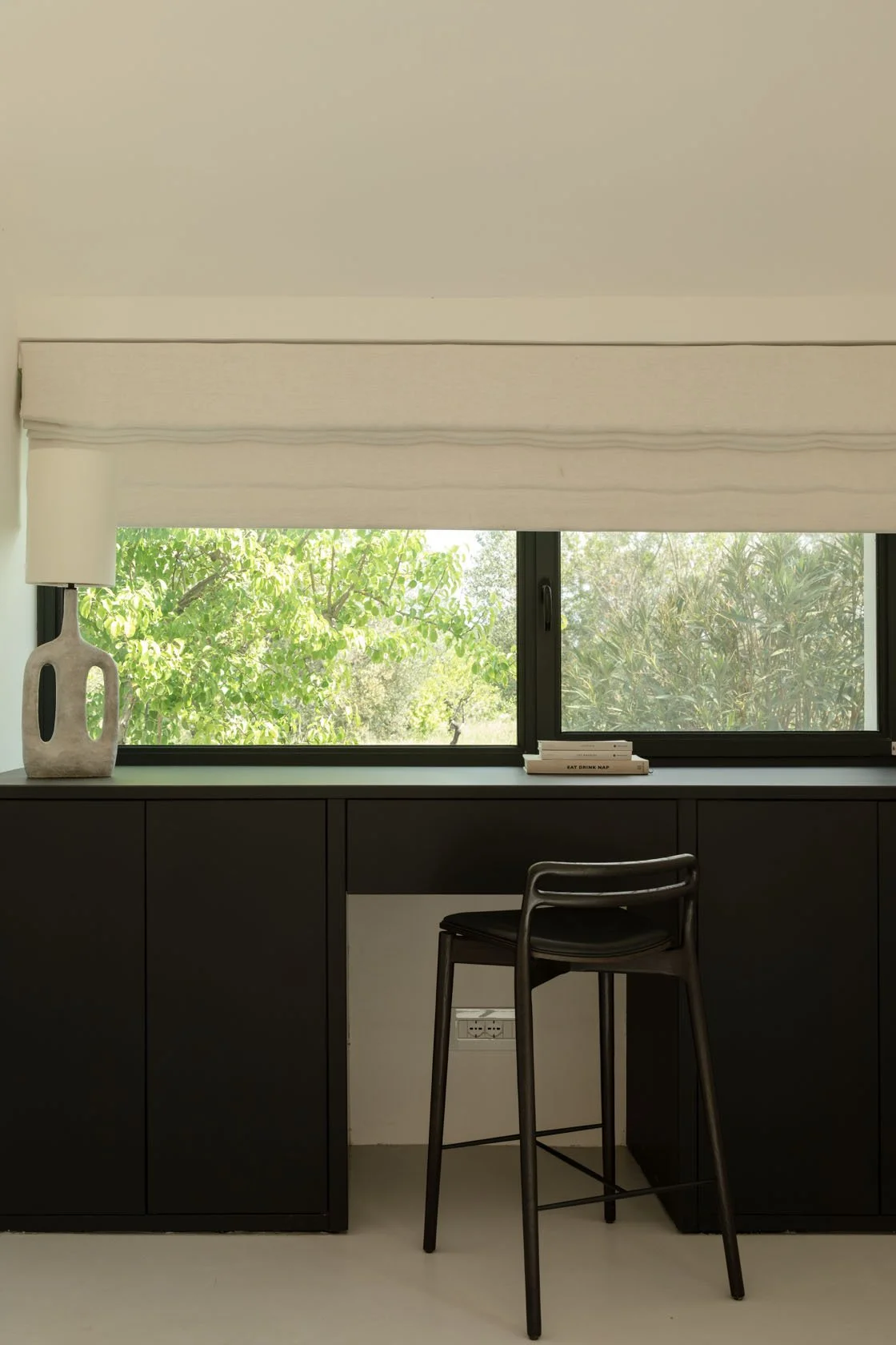 Interior view of a minimalist workspace with a black desk, a black chair, a window showing green foliage outside, a decorative lamp on the left, and a stack of books on the window sill.