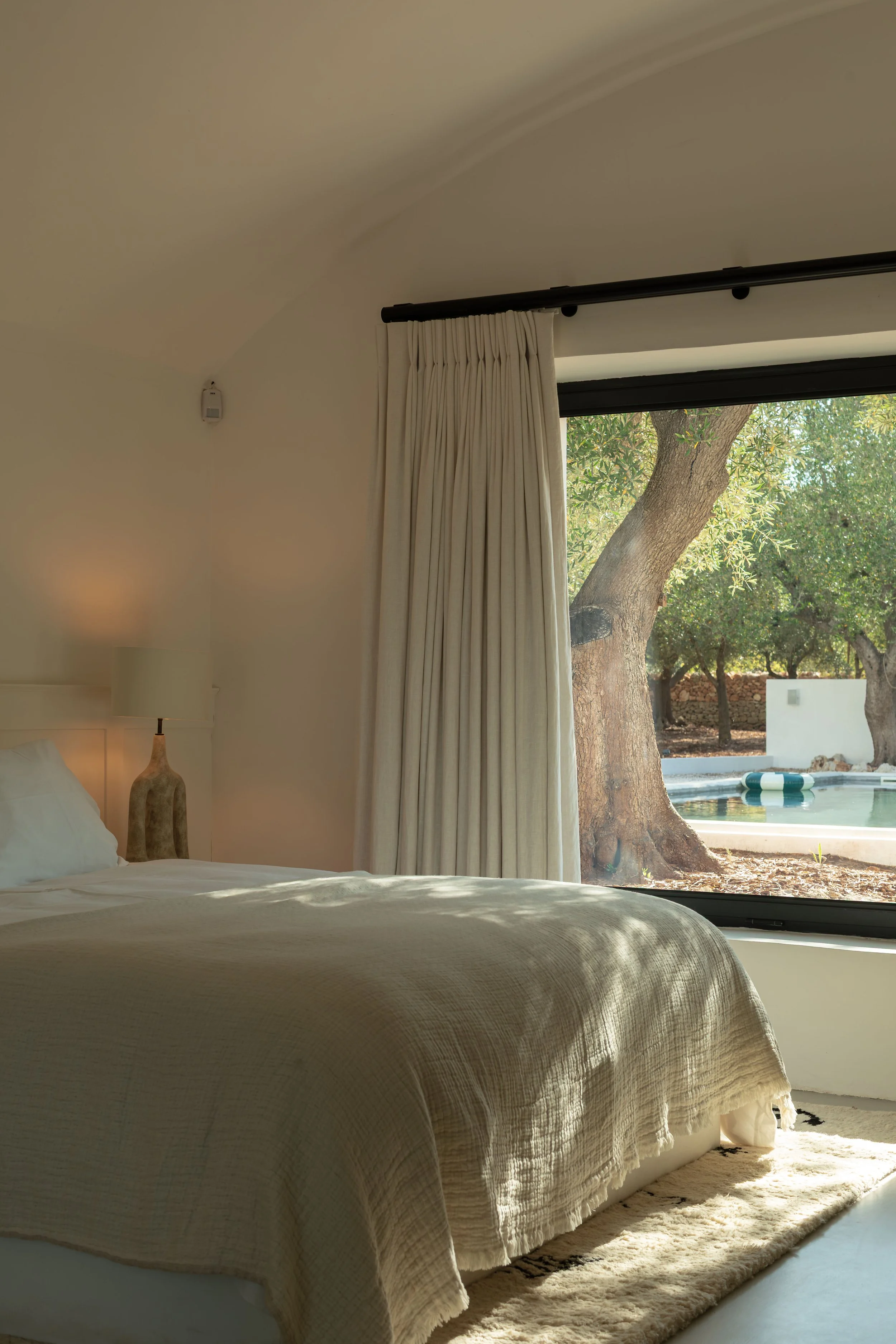 A bedroom with a large window showing a tree and an outdoor pool, a bed with beige linens, a nightstand with a beige lamp, and cream curtains.