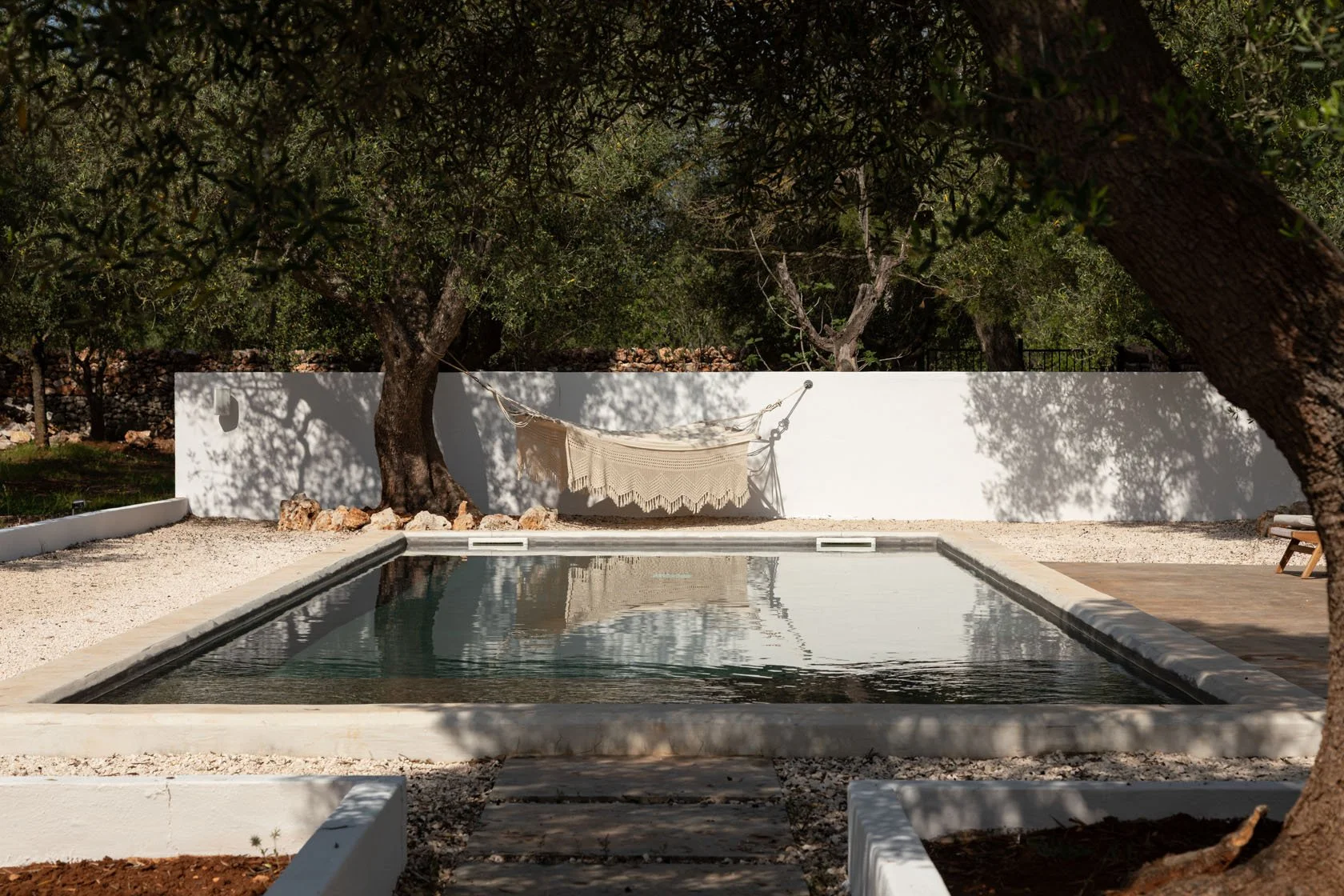 A backyard with a swimming pool surrounded by white gravel, trees, and a white wall. A hammock with a beige fringe blanket hangs between two trees, casting shadows onto the wall and pool area.