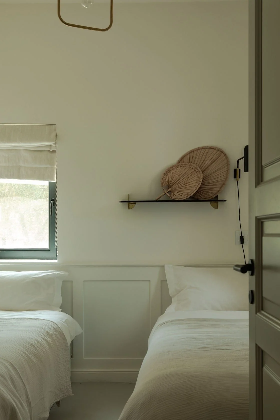 A cozy bedroom with two beds, a window with a beige shade, and a shelf on the wall with two decorative beige paper fans.