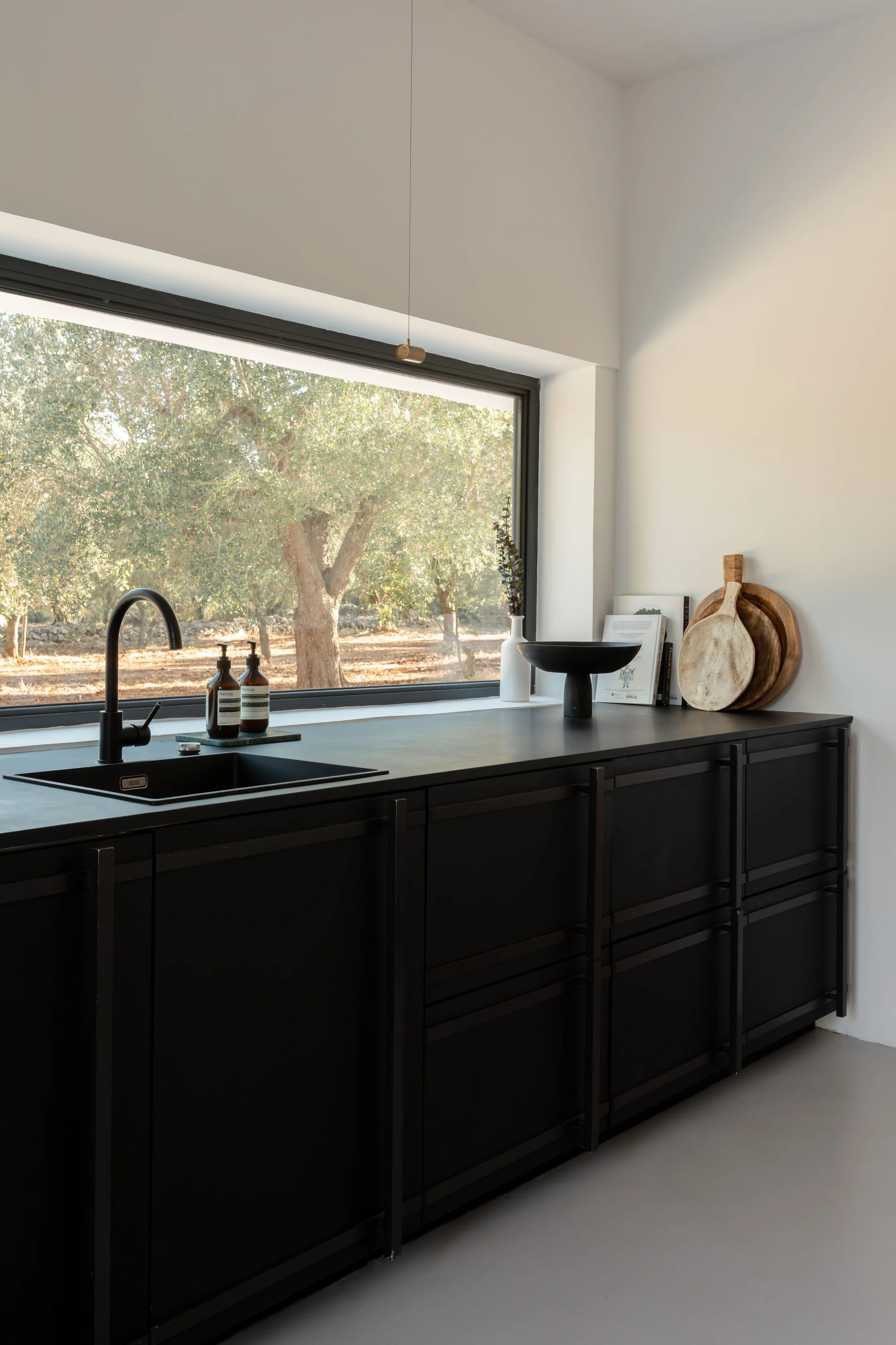 Modern kitchen with black cabinets, a large window overlooking trees, and minimalist decor including cutting boards, a bowl, and a vase.