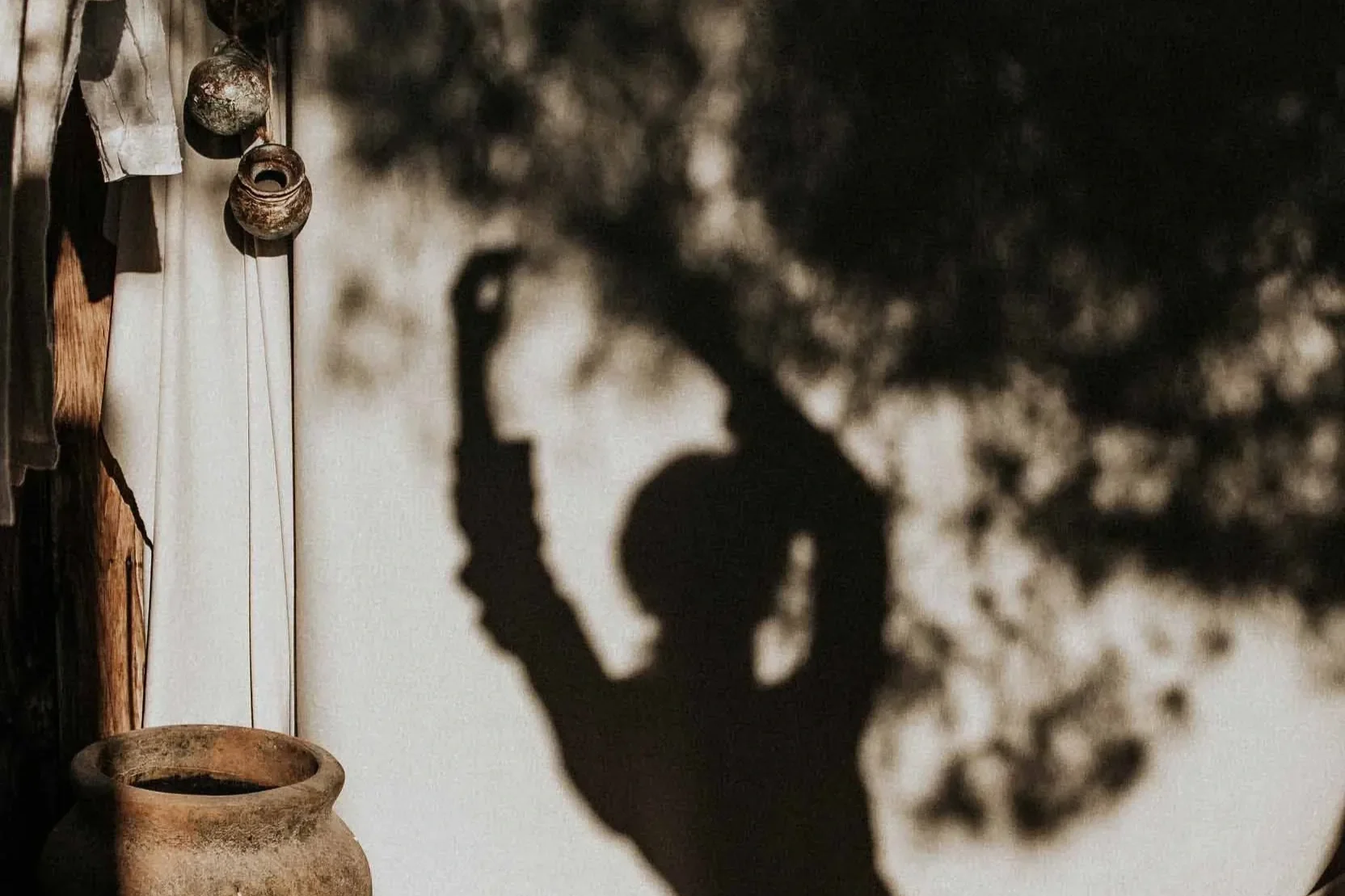 Shadow of a person with clay pots and hanging items on a wall, with sunlight casting the shadow and creating a silhouette effect.