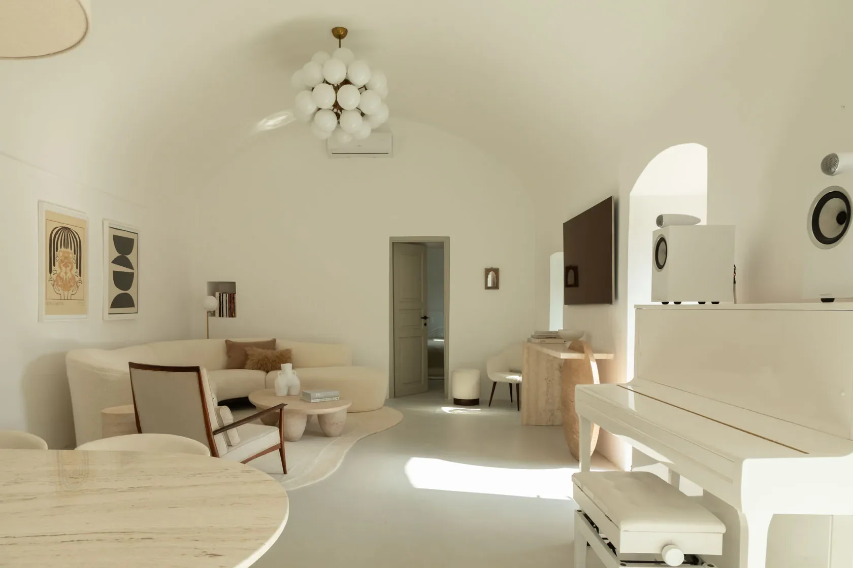 A minimalist living room with white walls, a cream-colored sofa with brown cushions, a modern coffee table, and abstract artwork on the wall. There's a white piano with speakers on top, and a large flat-screen TV mounted on the wall. The room features a spherical chandelier hanging from the curved ceiling, with natural light coming through a small arch window.
