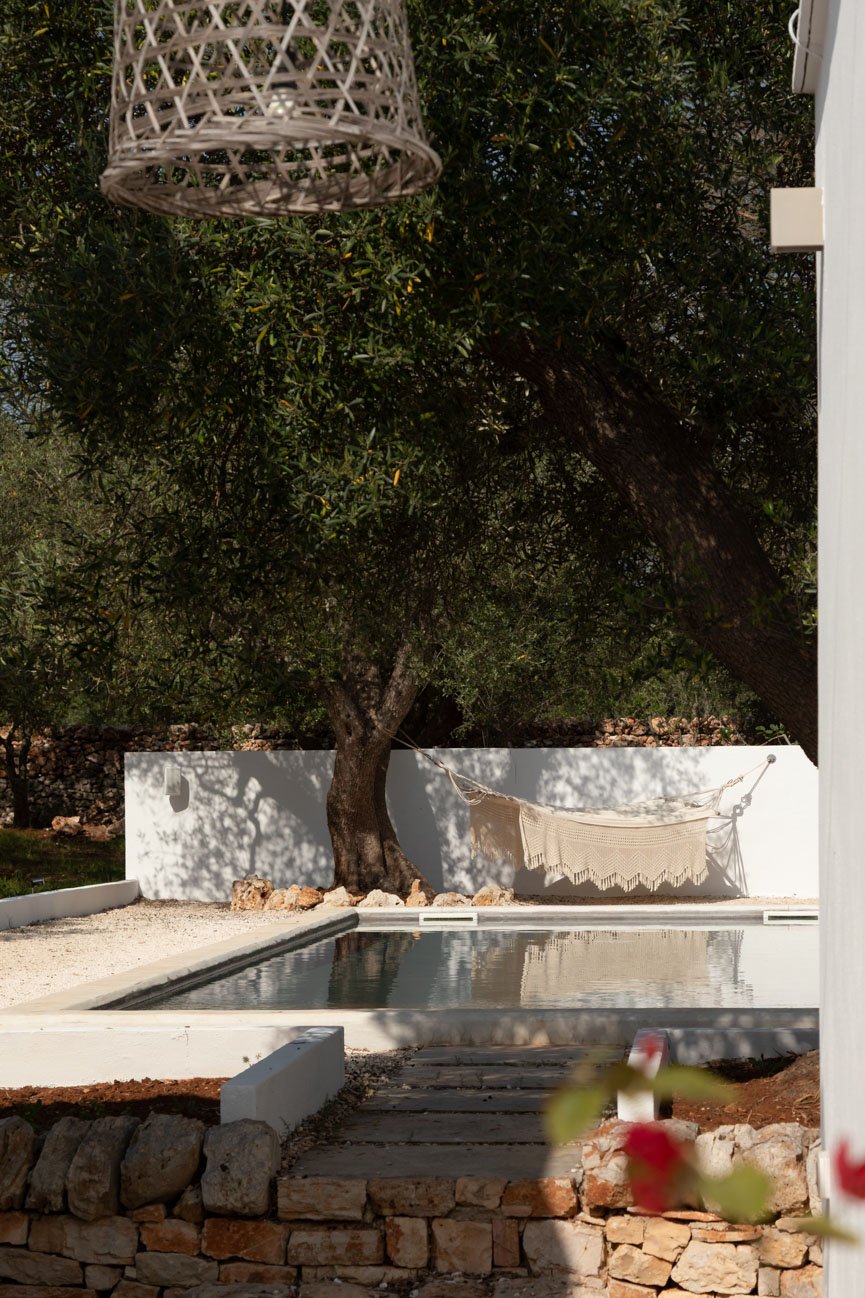 A backyard scene with a swimming pool, a large tree providing shade, a hammock hanging between the tree and a white wall, a stone-bordered pathway, and a wicker lampshade hanging overhead.