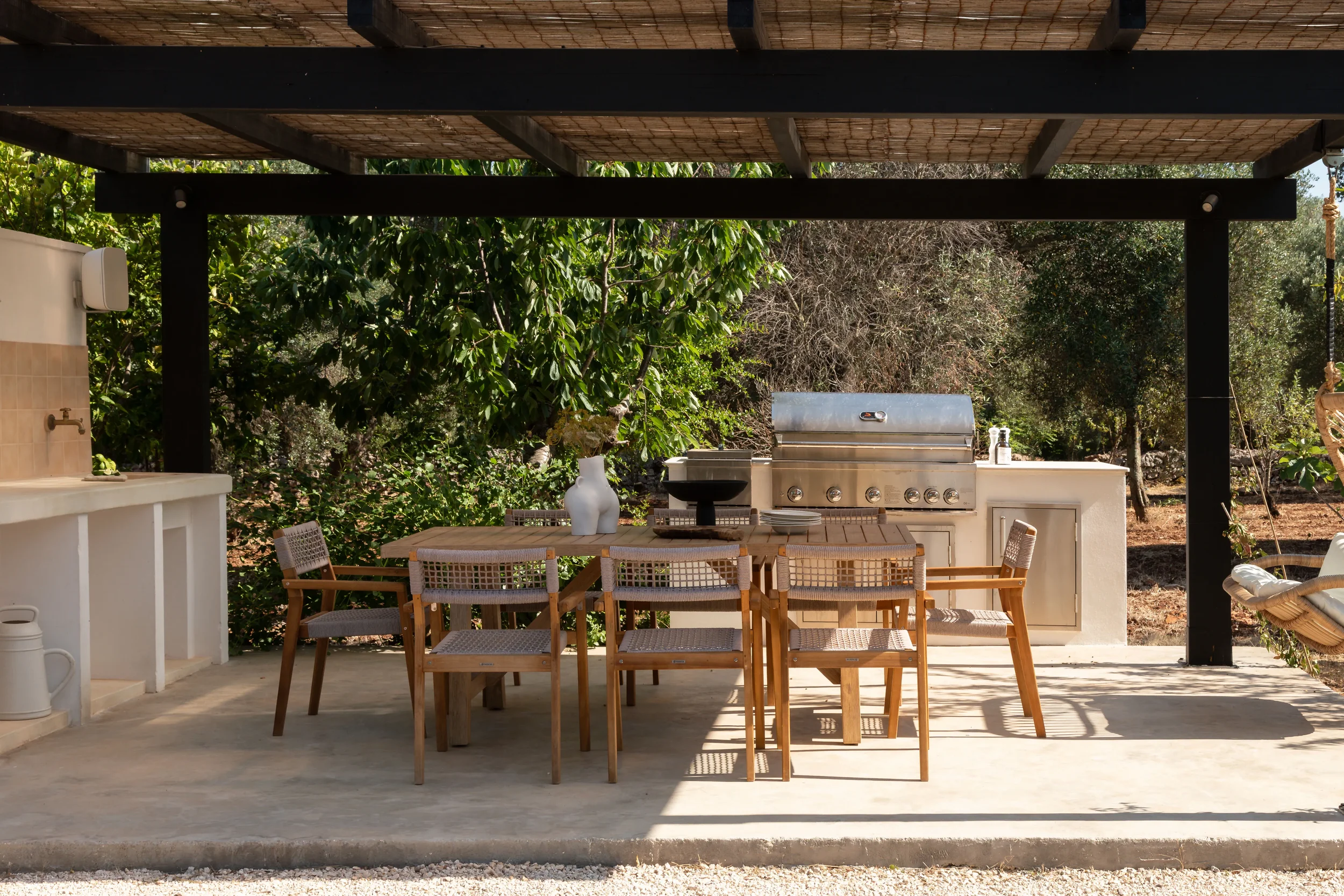 Outdoor patio area with a dining table, chairs, a built-in grill, and a sink, surrounded by trees and greenery.
