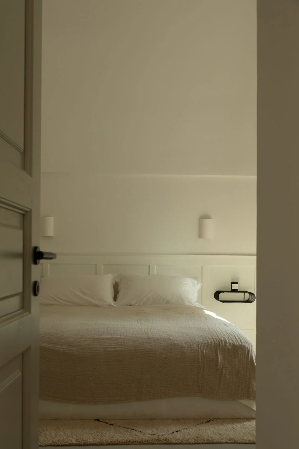 Photo of a neatly made bed with white pillows and beige bedspread, viewed through an open door in a minimalistic hotel room with white walls and wall-mounted light fixtures.