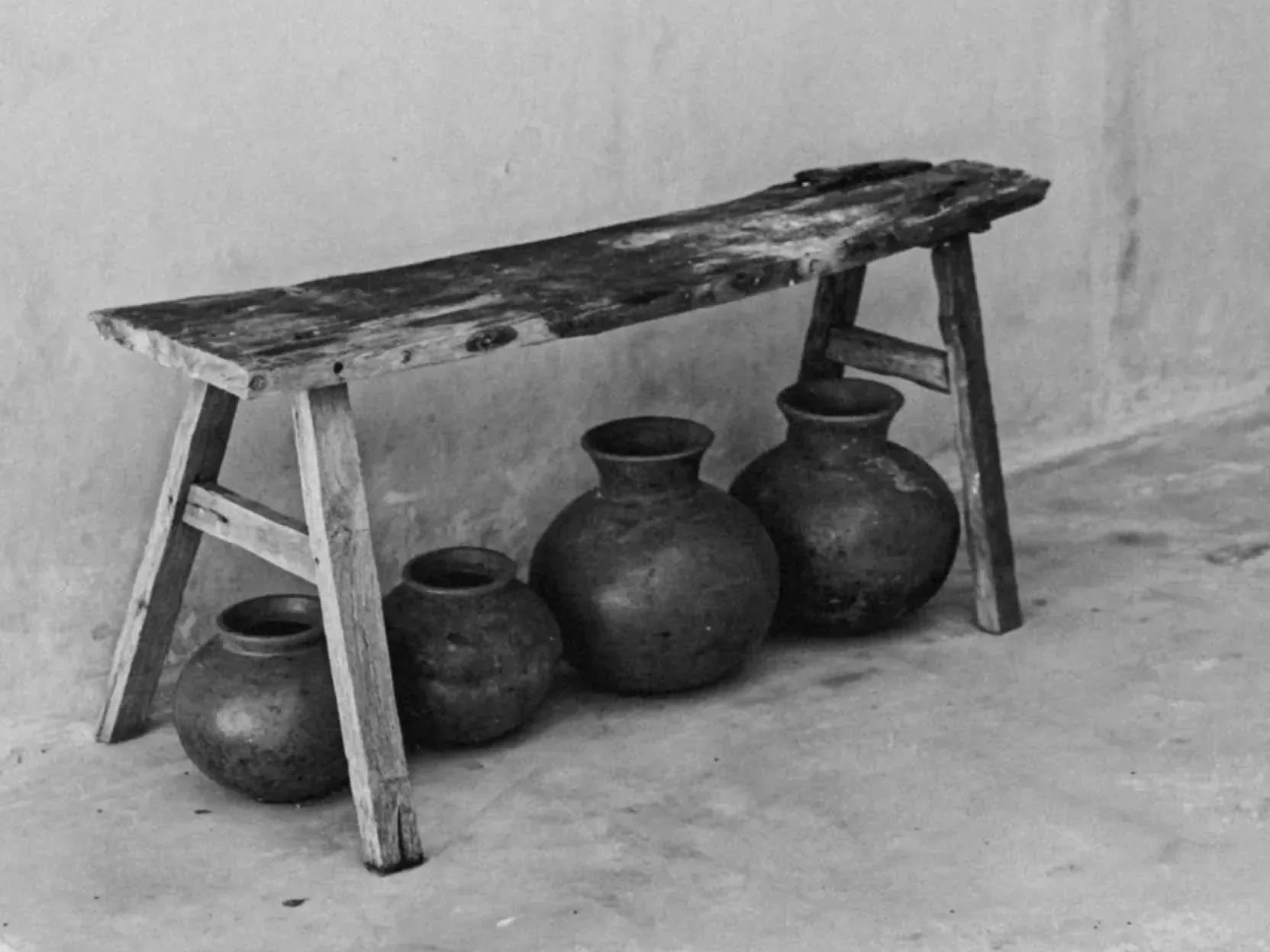 Black and white photo of a simple wooden shelf or bench with three large antique pots underneath.
