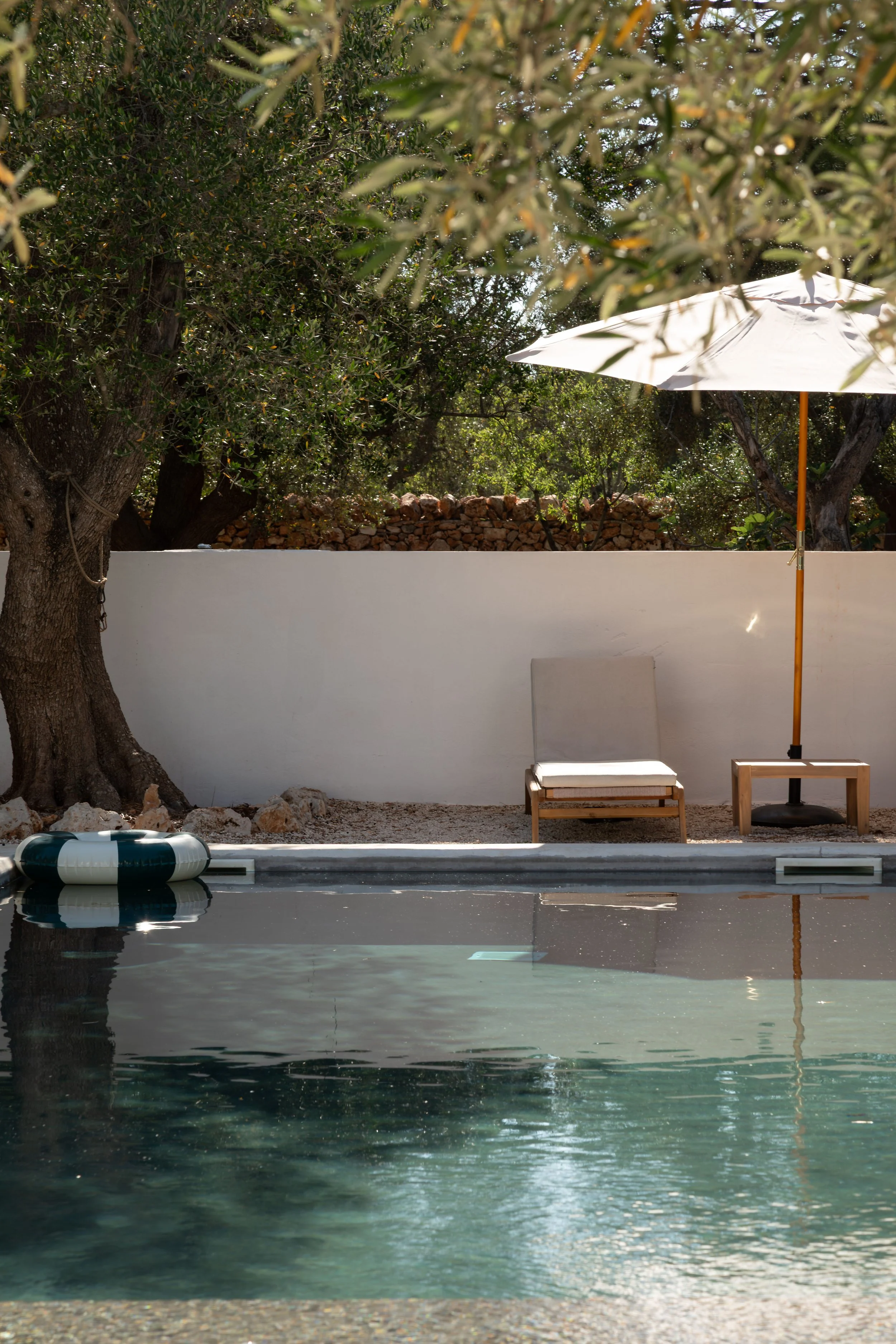 A backyard scene with a small swimming pool, a lounge chair, a white patio umbrella, and a white wall with trees behind it.