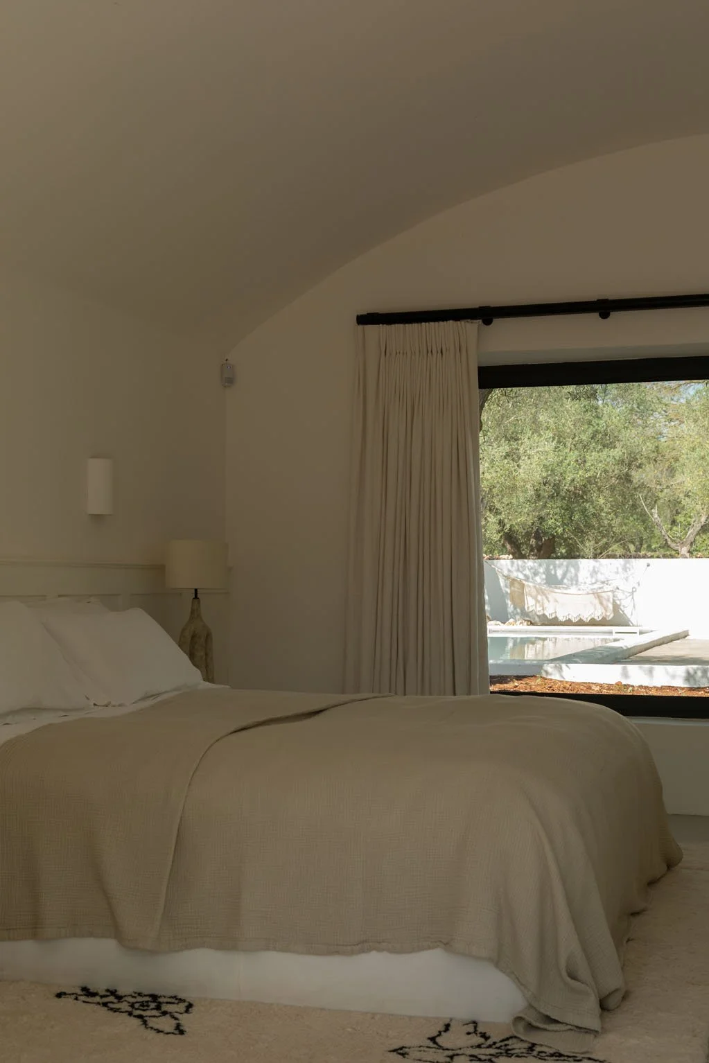 A bedroom with a beige bedspread, white pillows, and a nightstand with a lamp. There are cream-colored curtains and a large window showing green trees outside.