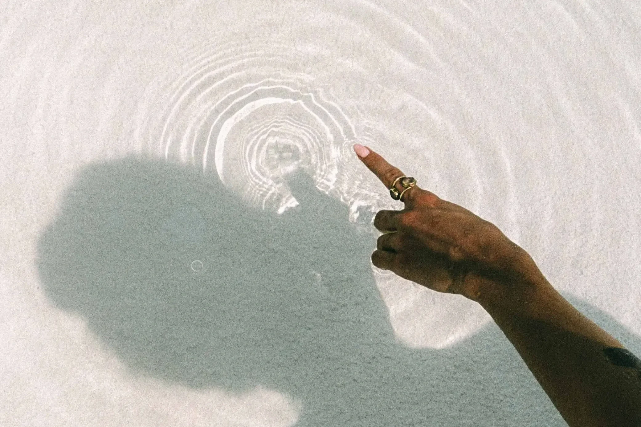 A person’s hand with rings touches the surface of white sand, creating ripples in the sand with their finger. The shadow of the person is cast on the sand.