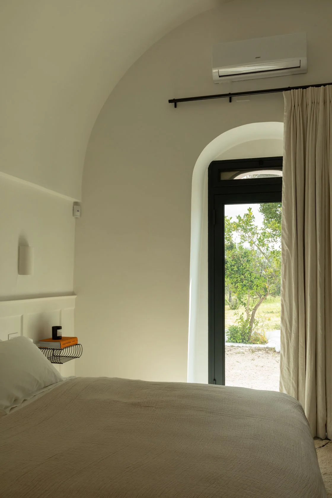 A bedroom with a bed, beige bedding, a wall-mounted lamp, a black window, cream curtains, an air conditioner, and a view of trees outside.