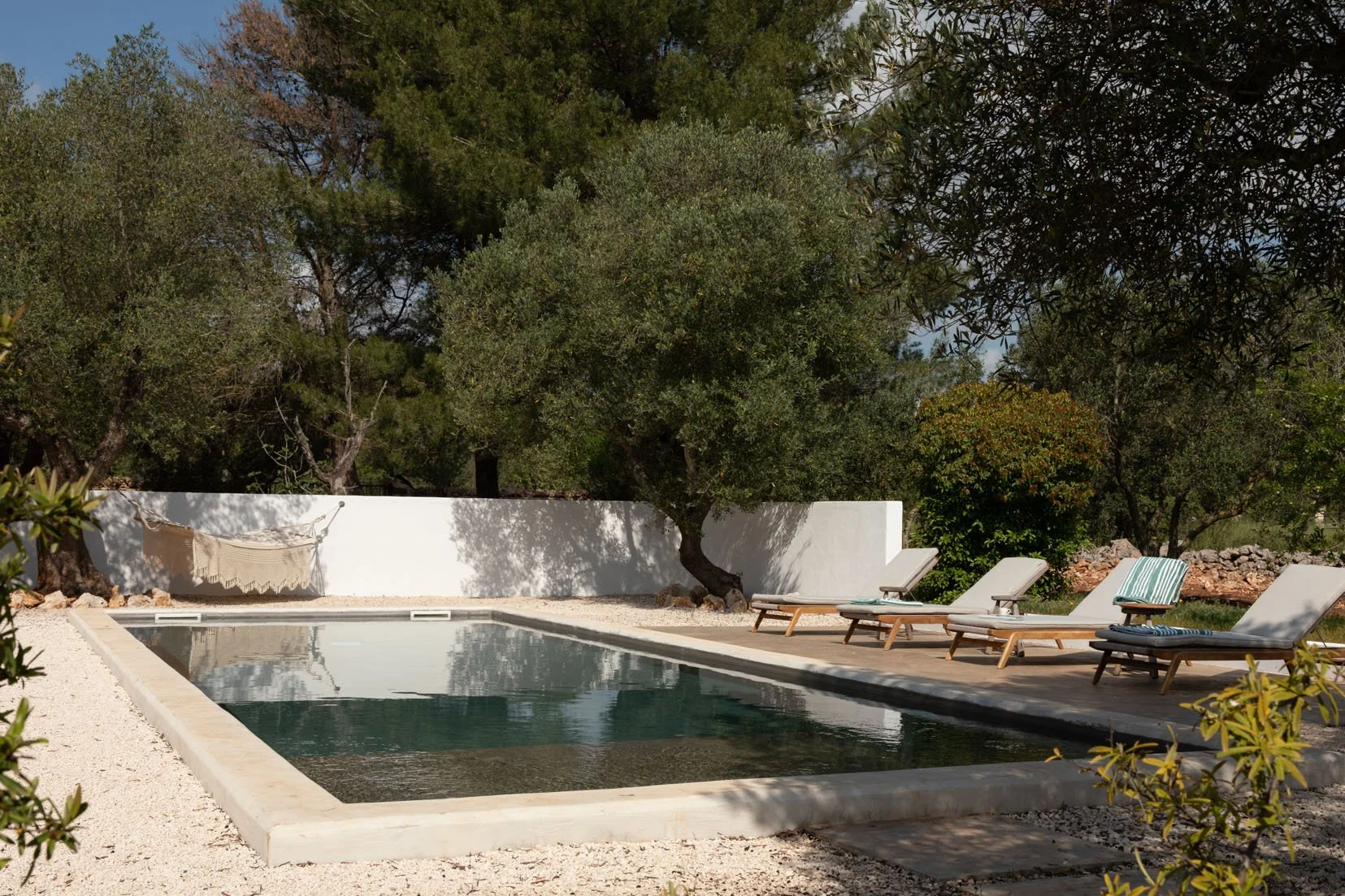 A swimming pool surrounded by lounge chairs and trees, with a white wall and a hammock in the background.