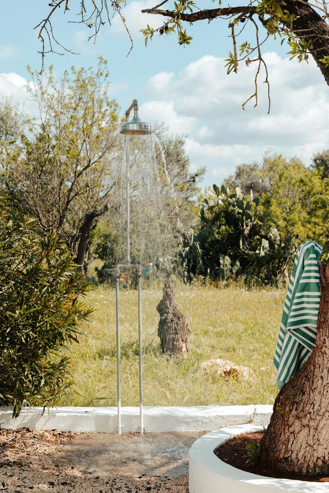 Outdoor shower with water running, surrounded by trees and greenery, with a striped towel hanging from a tree branch.