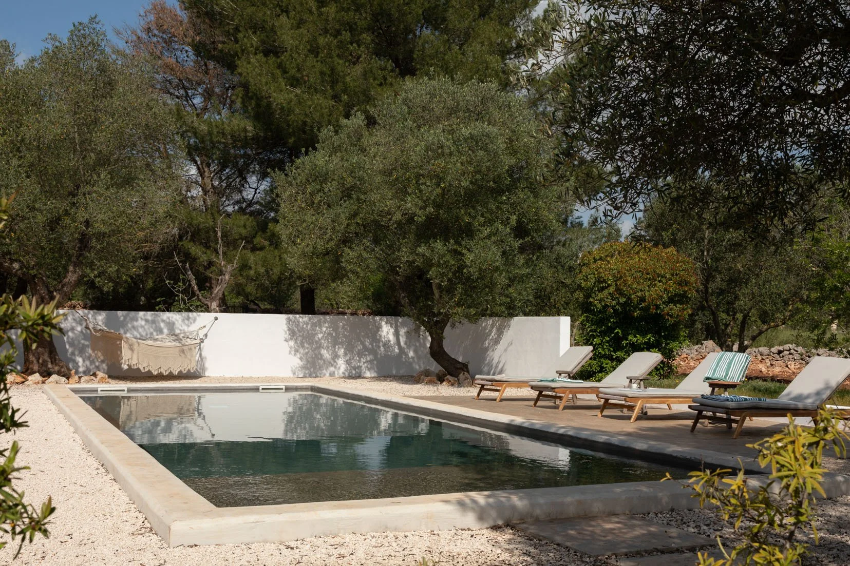 An outdoor swimming pool with deck chairs, a white wall, and trees in the background.