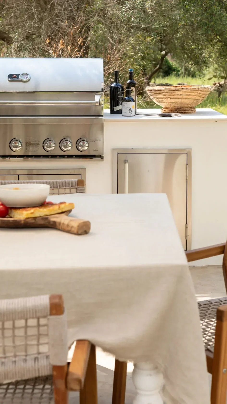 Outdoor kitchen with a stainless steel grill, four control knobs, bottles of wine and oil, a woven bowl on the counter, a table with a tablecloth, a pizza with toppings, a bowl of dip, and a chair in the foreground.