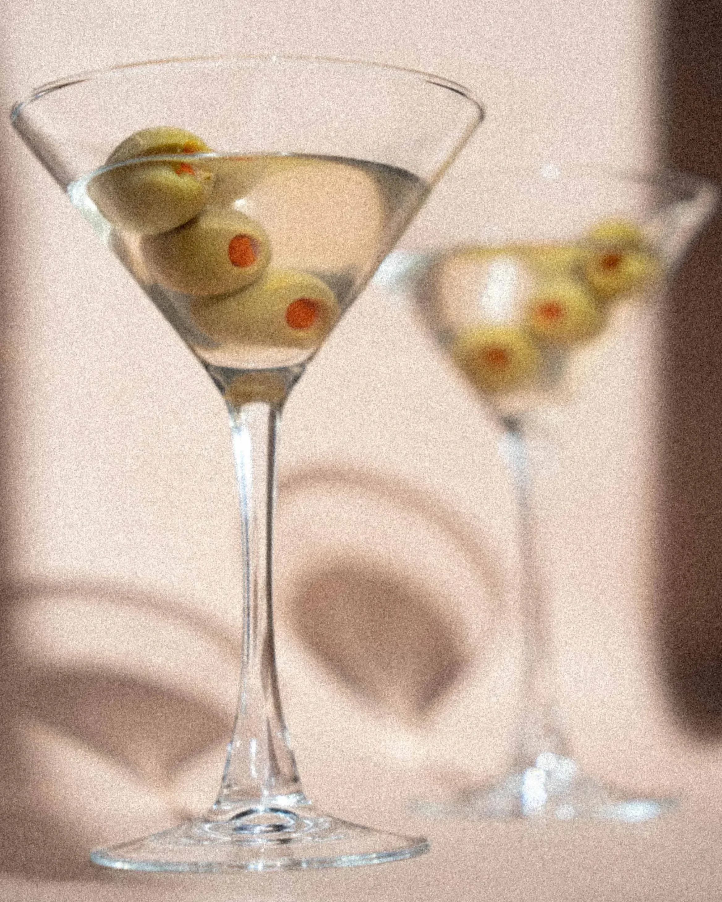 Two martini glasses with green olives served in a clear liquid, with a beige background and shadows cast on the surface.