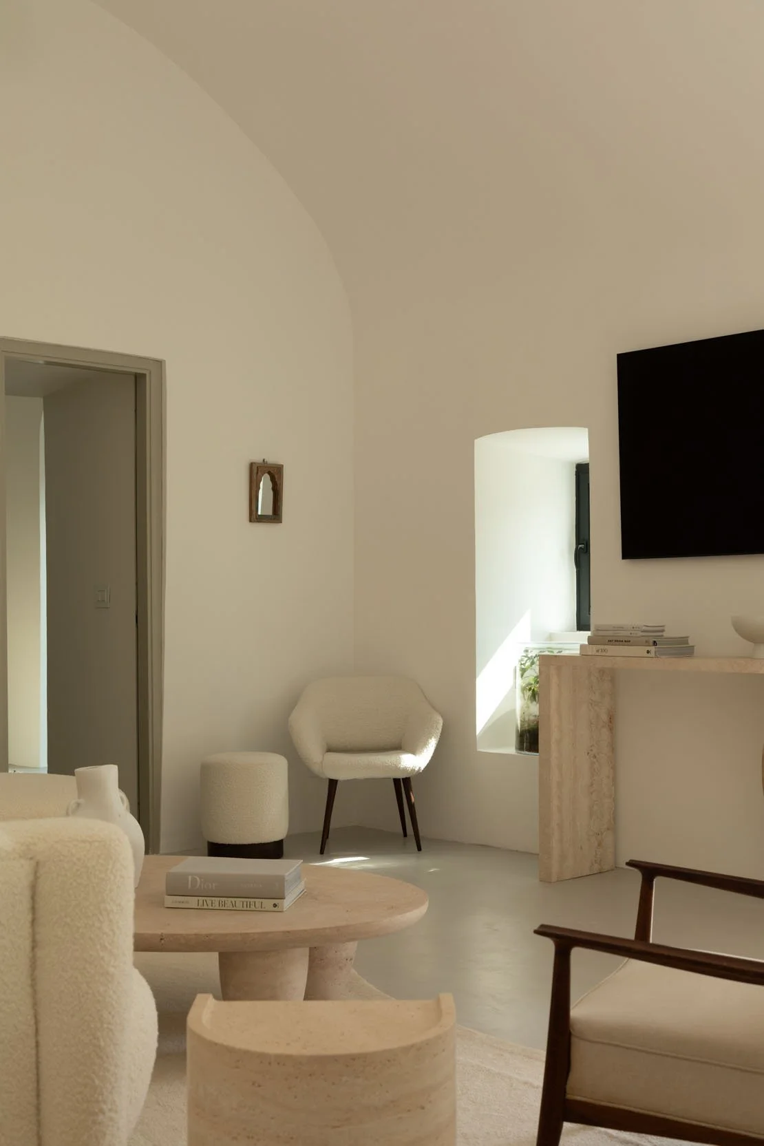 A minimalist living room with white walls and natural light. There is a small window, a wall-mounted TV, a white armchair, a beige round coffee table with books, and other light-colored furniture, creating a cozy and elegant atmosphere.