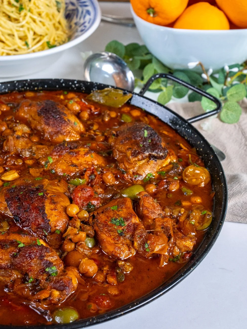 A pan of cooked chicken in tomato-based sauce with chickpeas and vegetables, garnished with herbs. In the background, bowls of spaghetti and oranges.