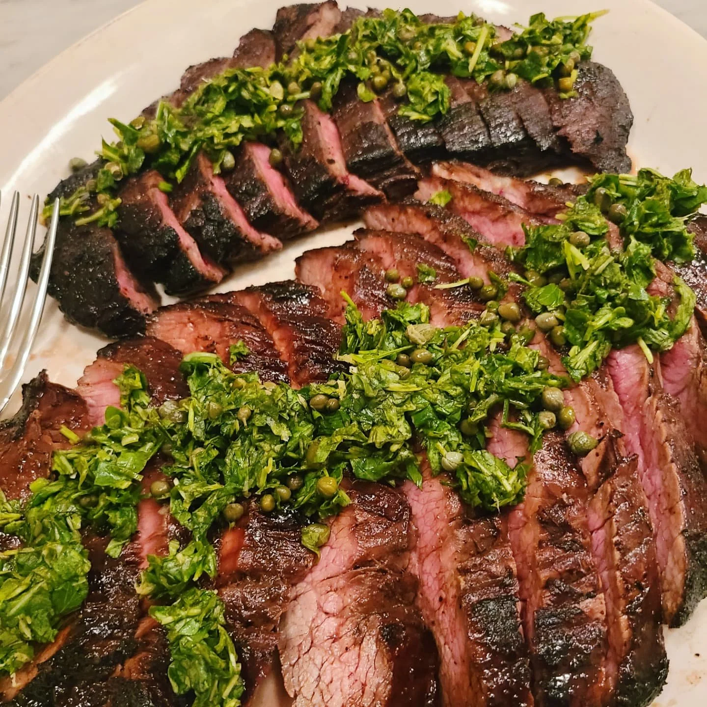 Sliced cooked steak topped with chopped green herbs and peppercorns on a white plate.