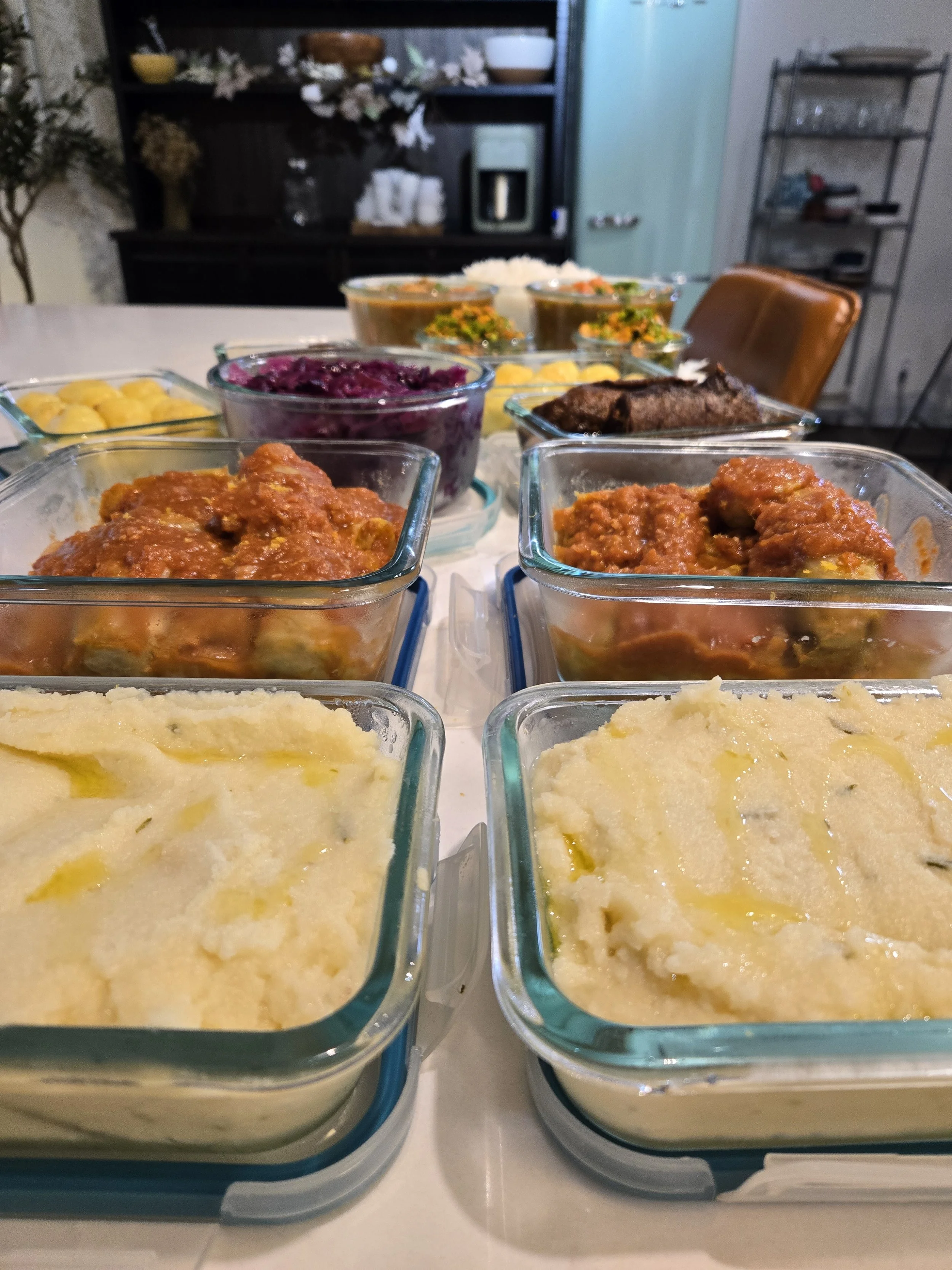 A variety of dishes in glass containers on a table, including mashed potatoes with butter, meat in sauce, purple cabbage, and other side dishes in bowls, with a kitchen background.