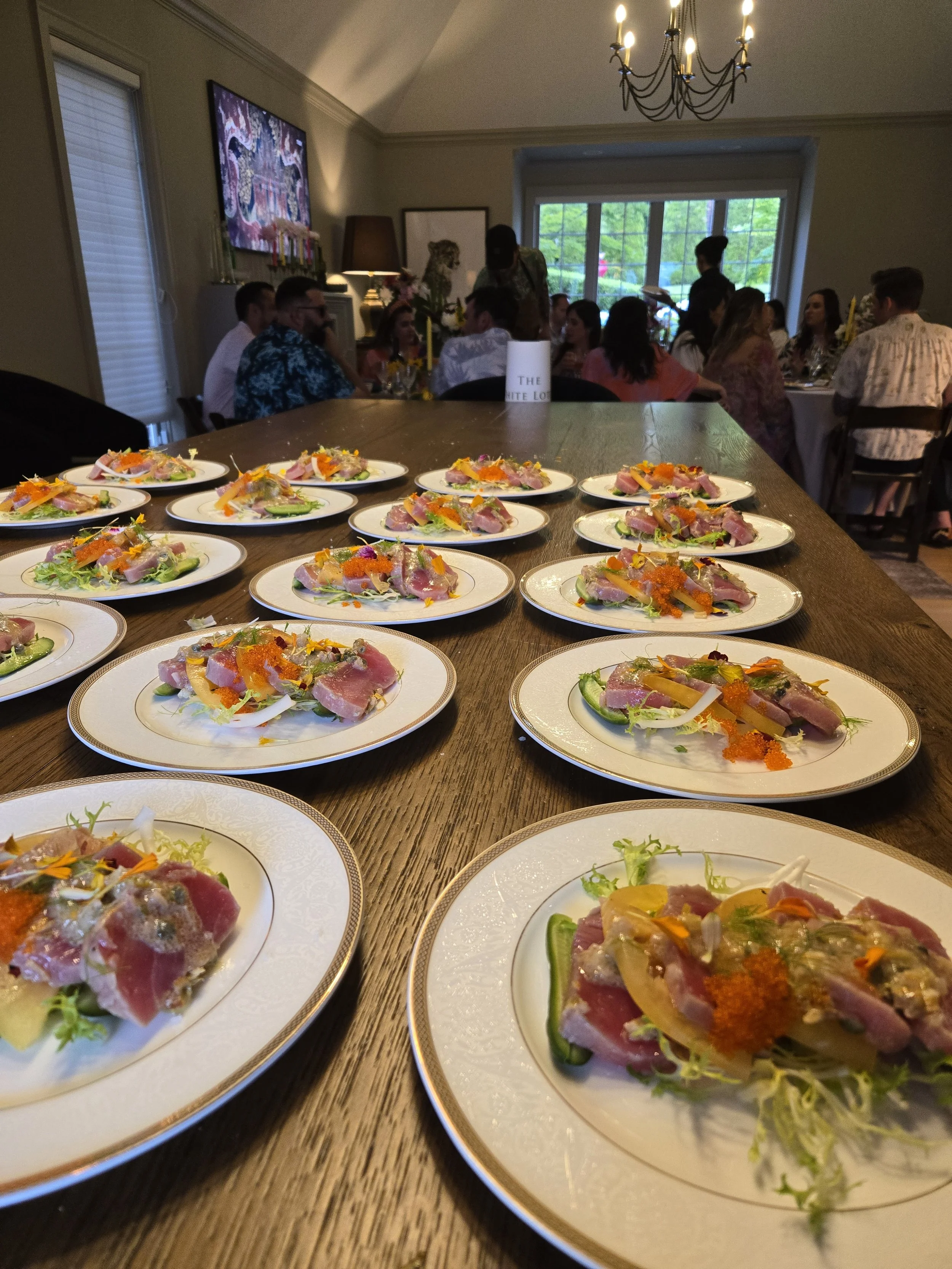 Multiple plates of colorful sashimi or seafood salads arranged on a long wooden table in a well-lit dining room with people seated at tables in the background.