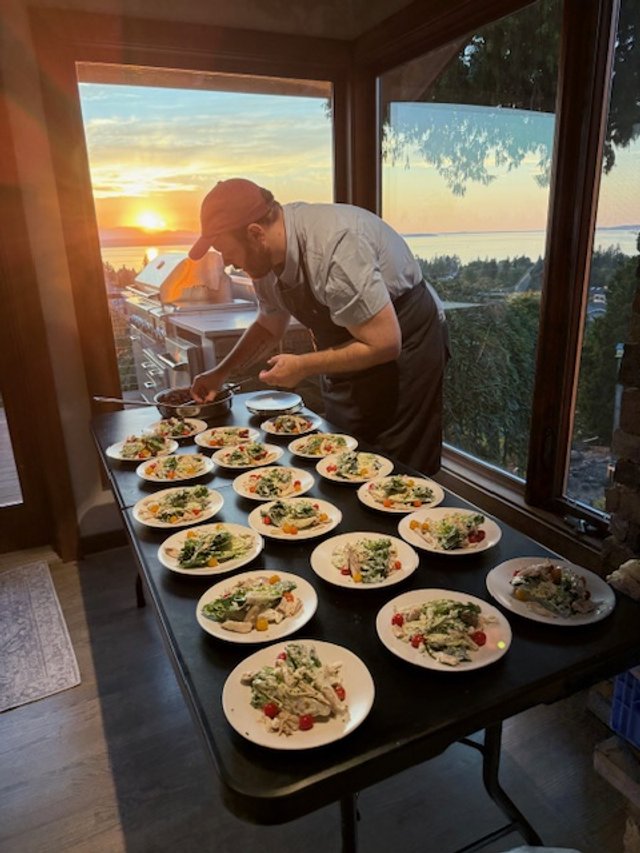 A chef prepares multiple plates of food on a table by a large window at sunset, with a scenic view of trees, water, and the sky.