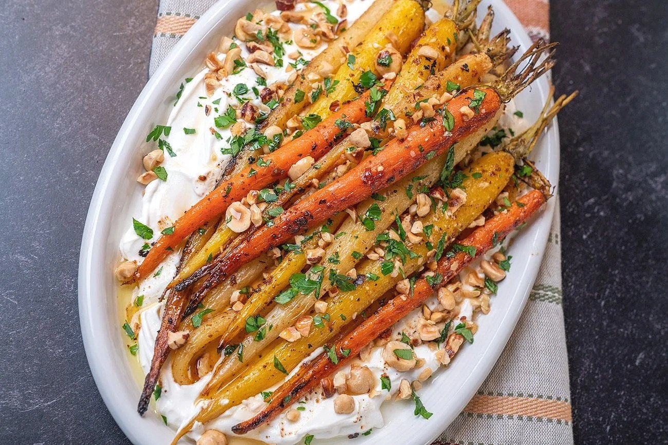 A white oval dish filled with roasted rainbow carrots on a bed of creamy white sauce, garnished with chopped herbs and crushed nuts.