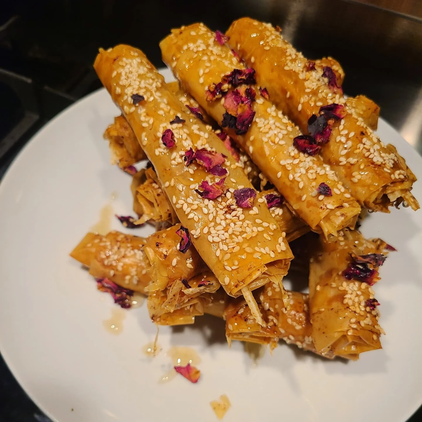 Stack of golden-brown phyllo rolls sprinkled with sesame seeds and dried rose petals on a white plate.