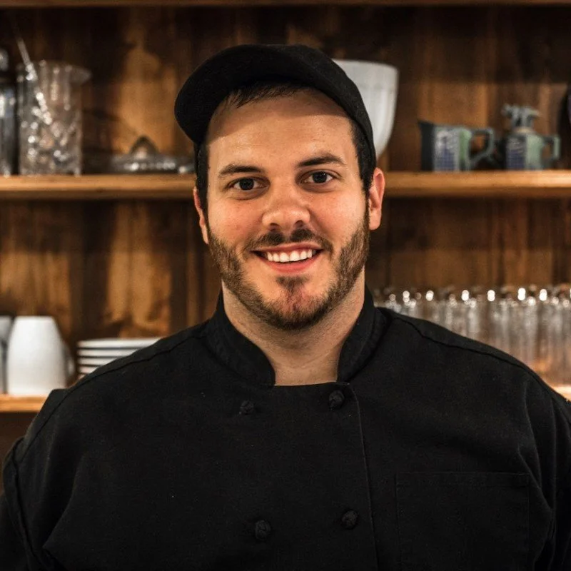 Chef owner Dom Ruane , a smiling man in a black chef's coat and cap standing in front of a wooden shelf with glasses and other kitchen items.