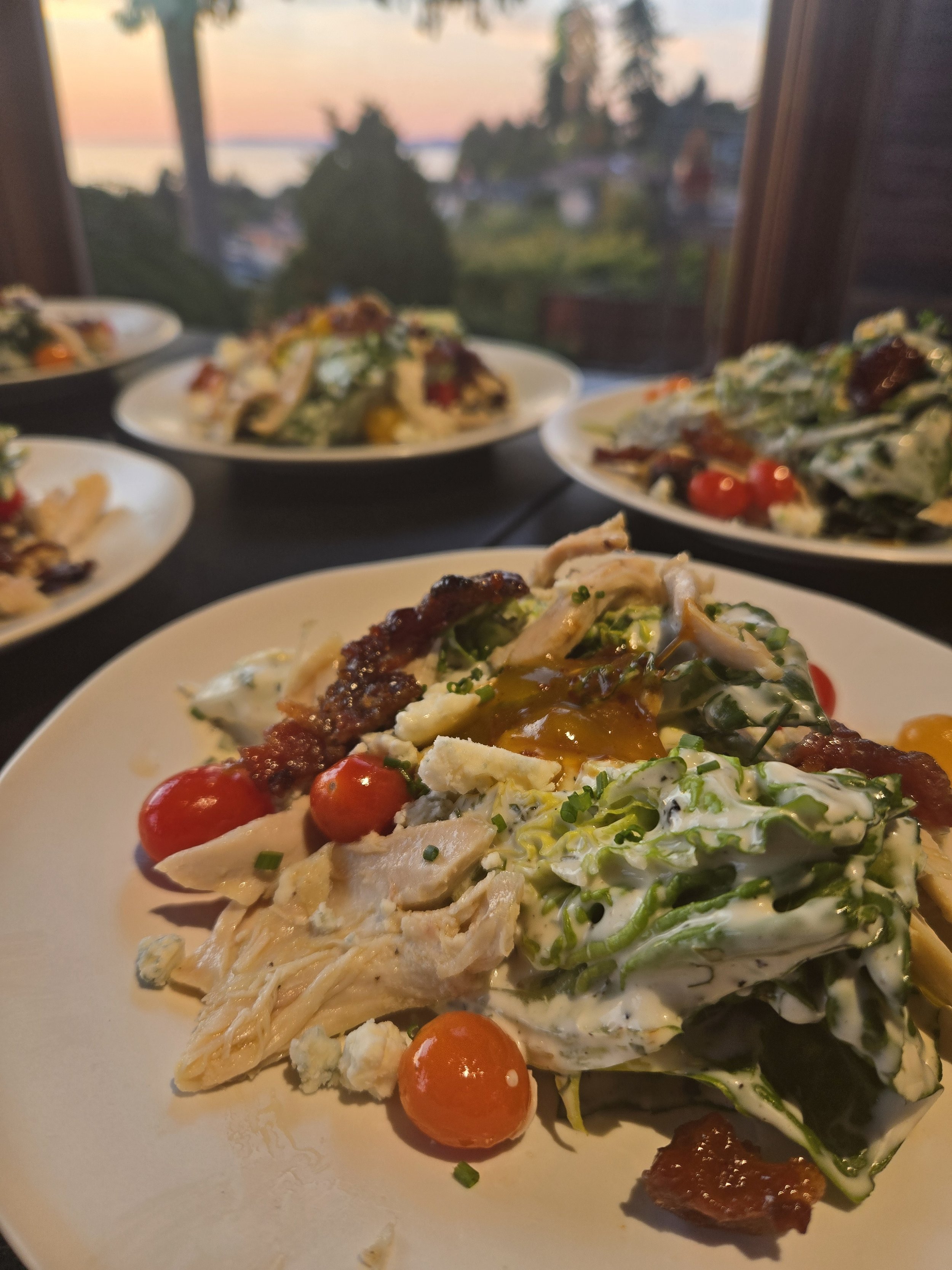 Salad with cherry tomatoes, chicken, bacon, and creamy dressing served on a white plate with a sunset view in the background.