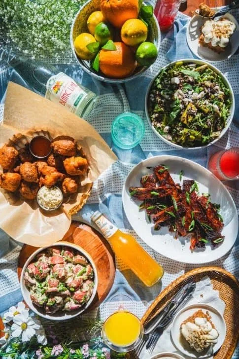 A table with various foods including a bowl of colorful fresh fruits, a bowl of salad greens, a plate of chicken wings with dipping sauces, a bowl of pasta salad, a plate of glazed ribs garnished with herbs, and beverages like orange juice and other drinks.