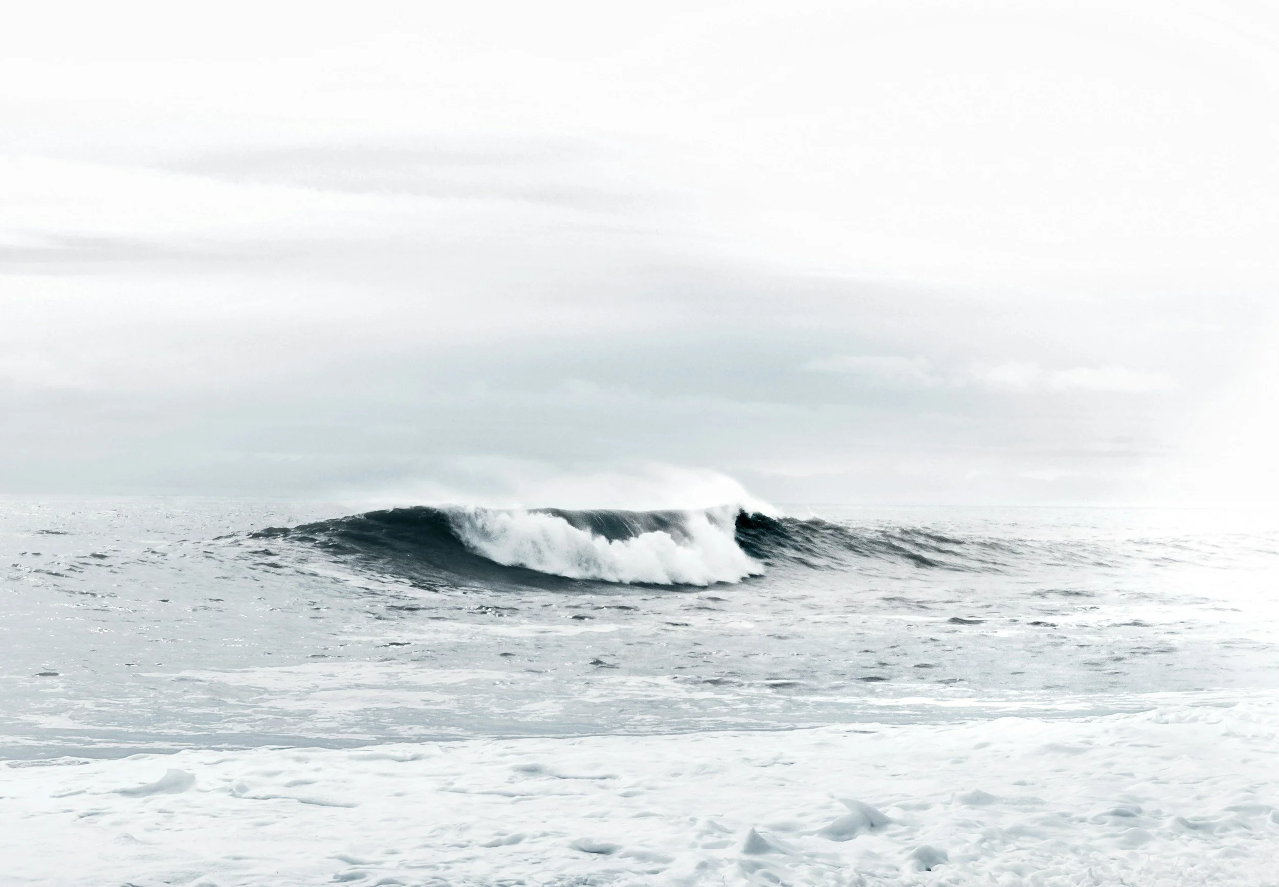 Ocean waves crashing on a snow-covered shoreline under a cloudy sky.