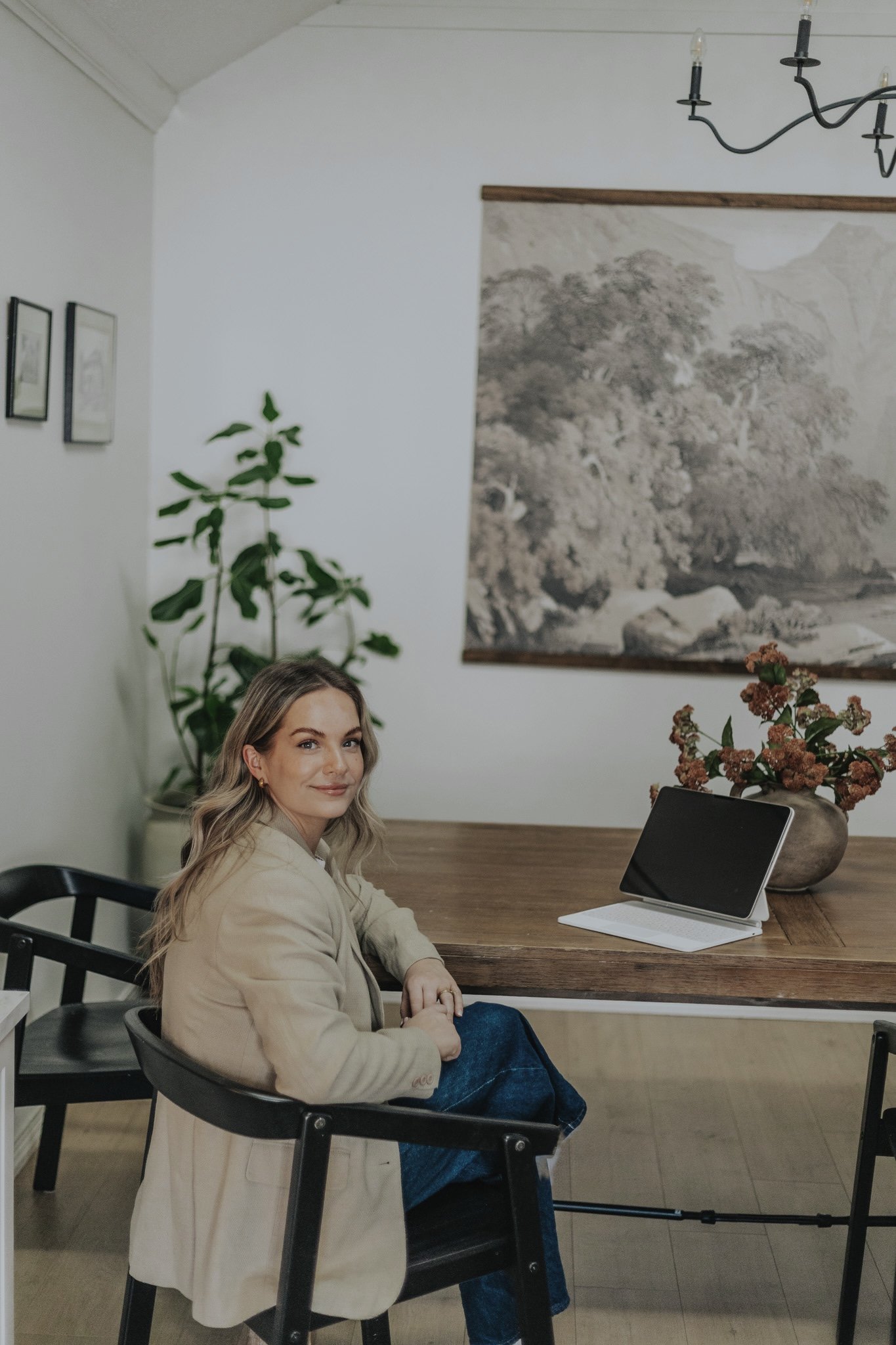 A woman sitting at a wooden table in a home or office setting, with a tablet and a vase of flowers in front of her. There are framed pictures on the wall and a large plant in the background.